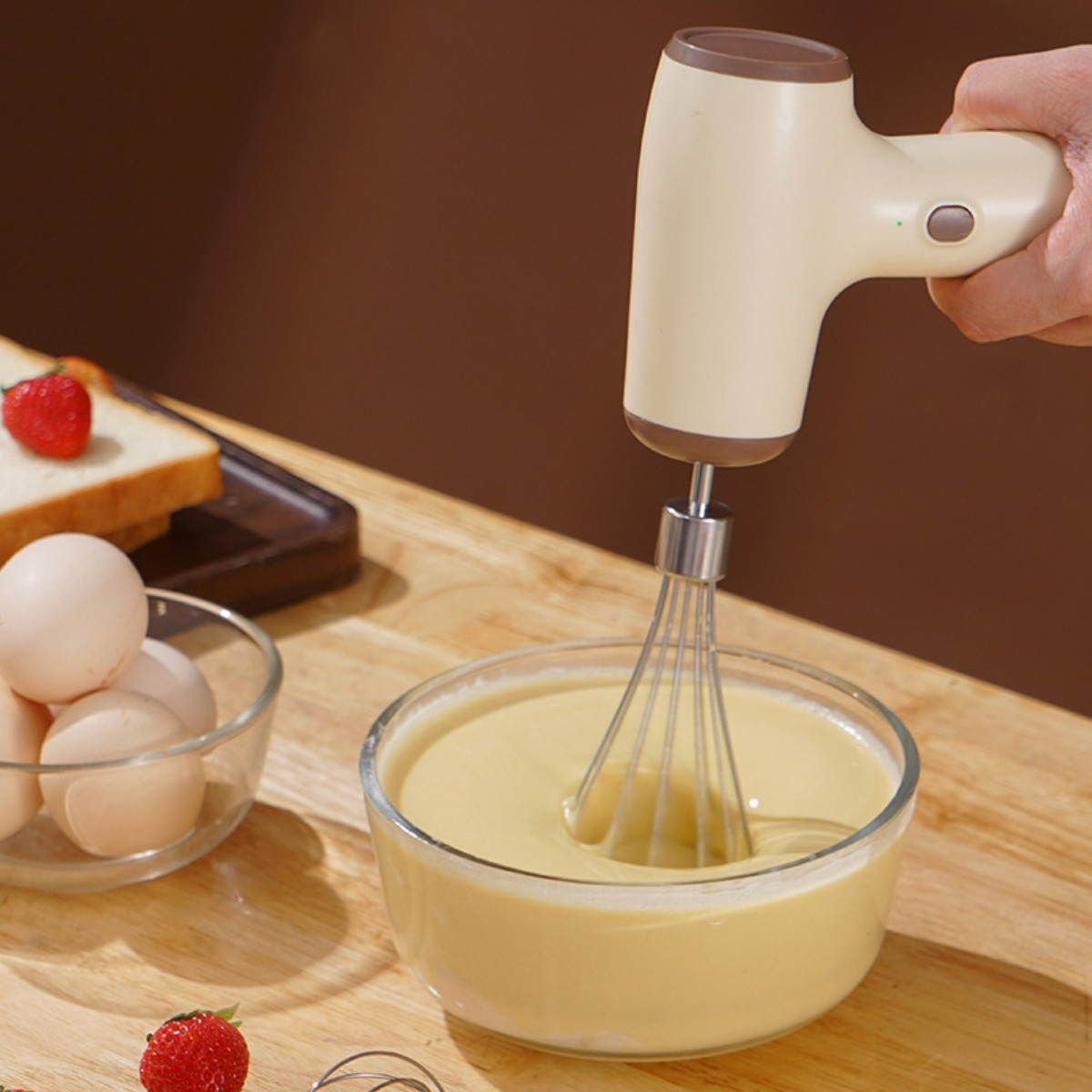 Handmixer verrührt Teig in Glasschüssel. Eier, Erdbeeren und Brot liegen daneben.