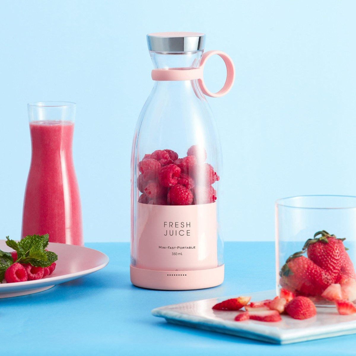 Licuadora portátil rosa con bayas dentro, junto a un vaso con jugo rosa.