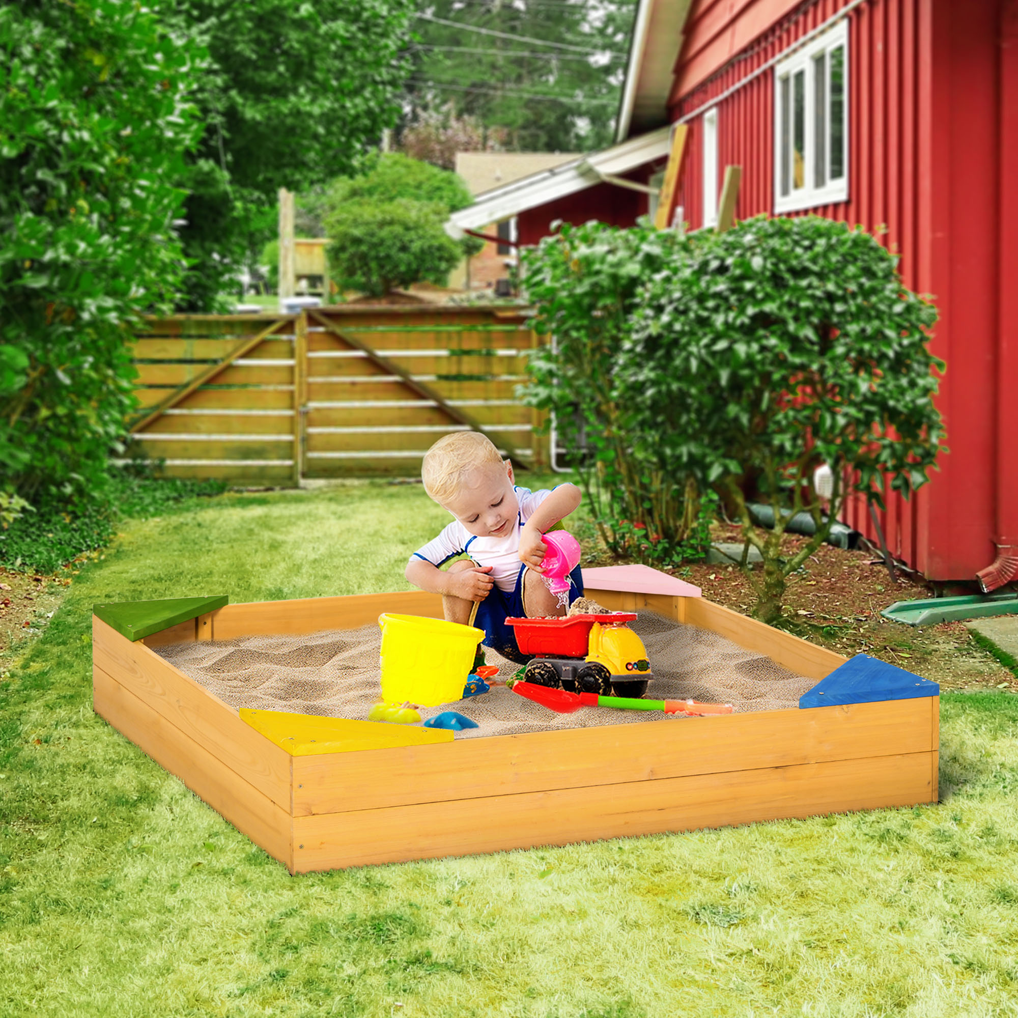 Un niño jugando en un arenero de madera con arena, juguetes y una casa roja al fondo.