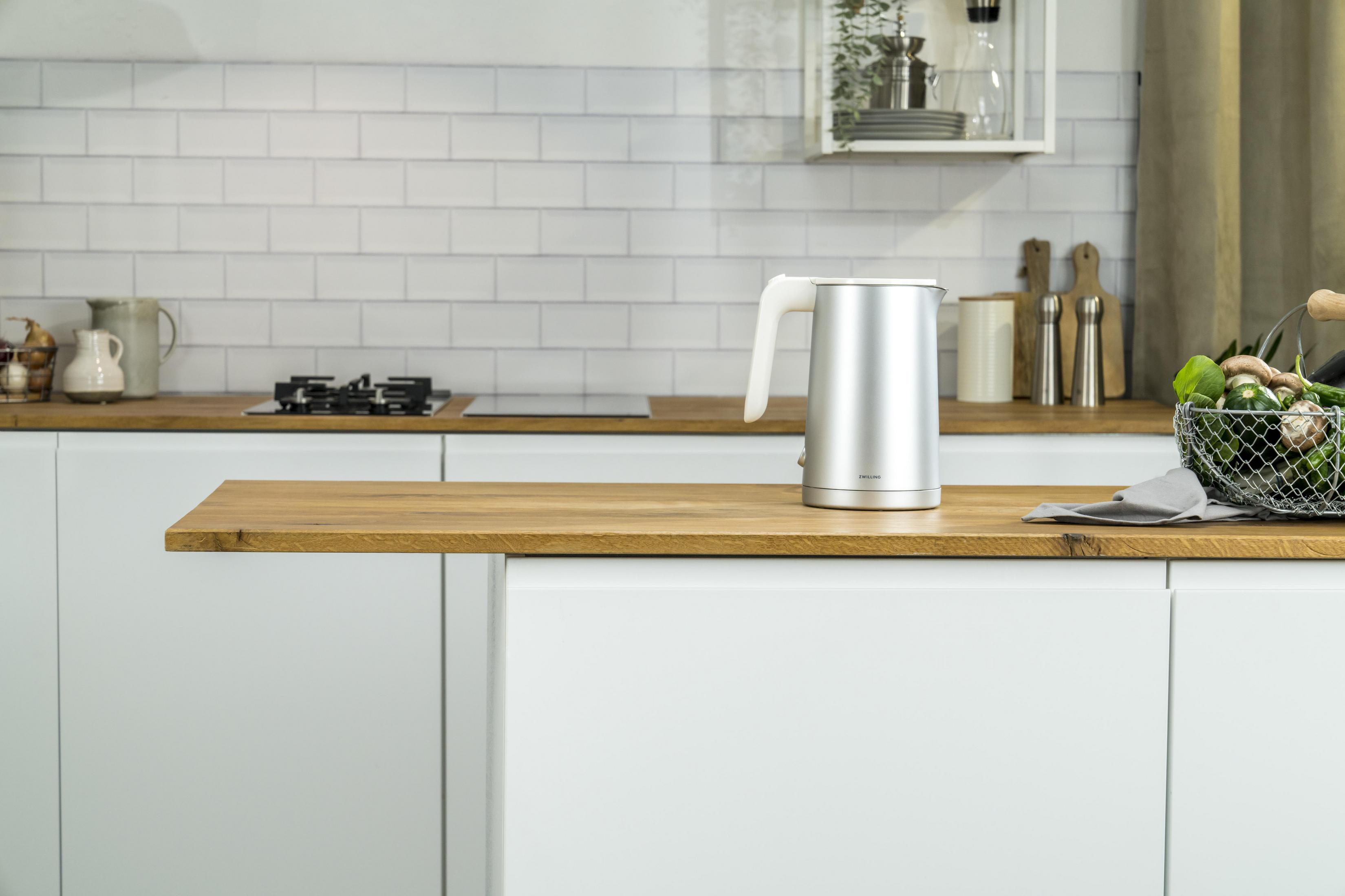 Nowoczesna kuchnia ze srebrnym czajnikiem elektrycznym na drewnianym blacie i białymi szafkami.