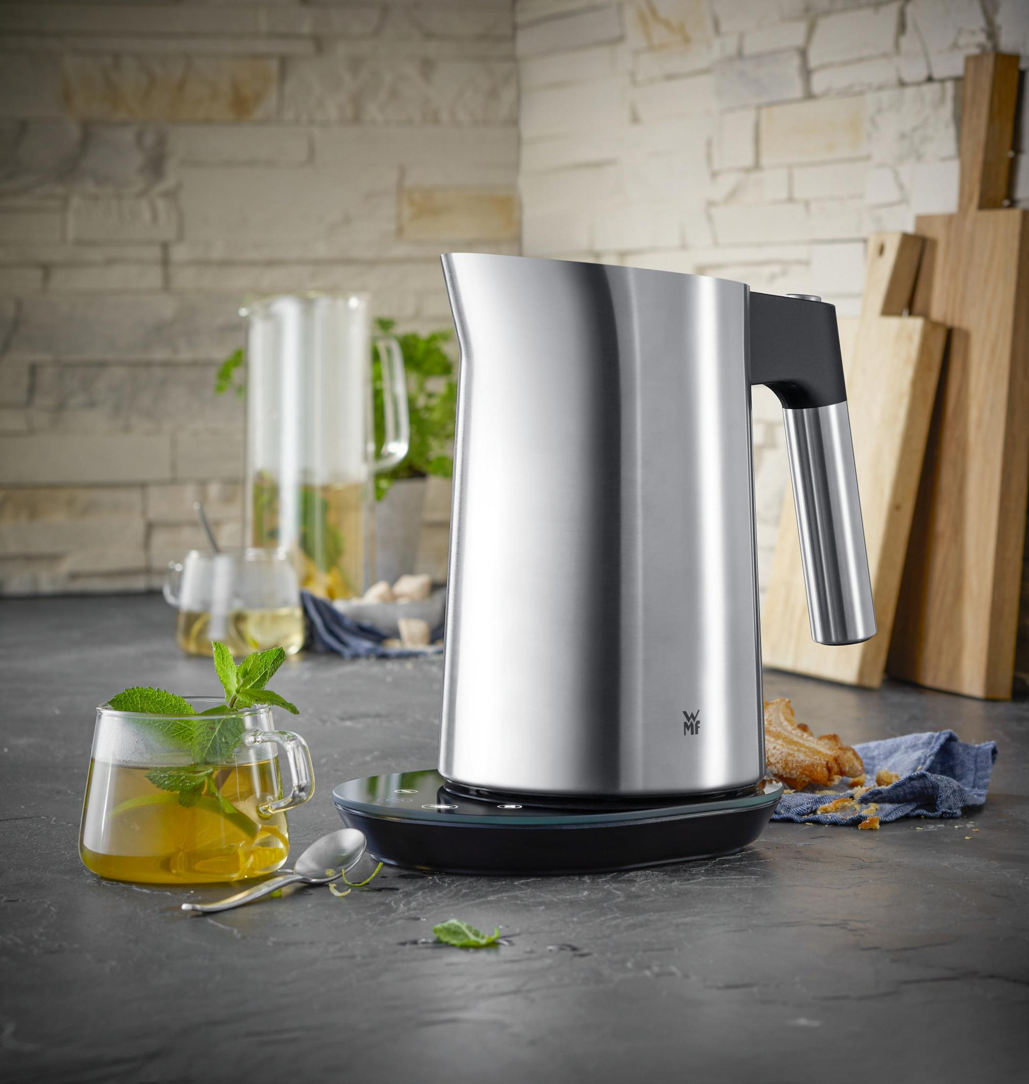 Srebrny czajnik na czarnej podstawie. Szklanka herbaty i jedzenie w tle.