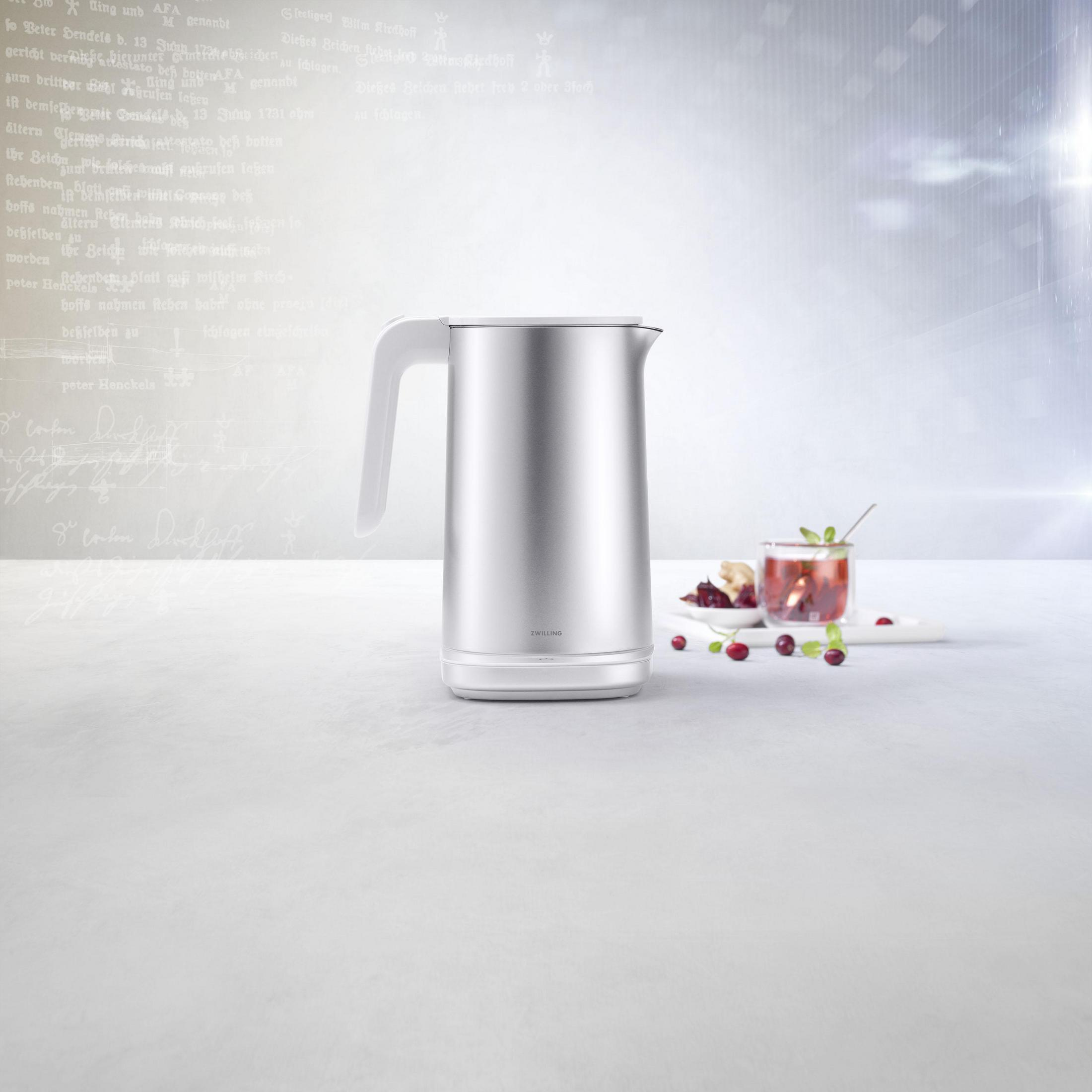 Srebrny czajnik elektryczny z białym uchwytem. Talerz z jagodami i filiżankami herbaty w tle.