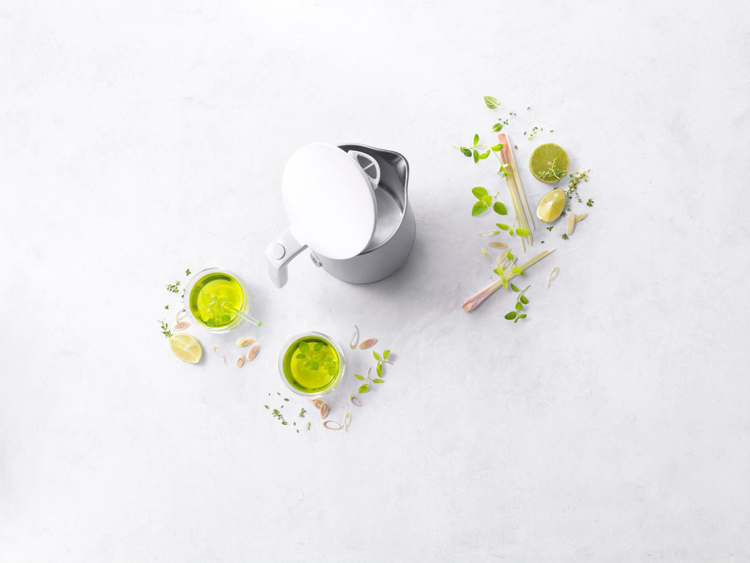Zaparzacz do herbaty z pokrywką, dwie szklanki herbaty i zioła na białej powierzchni.