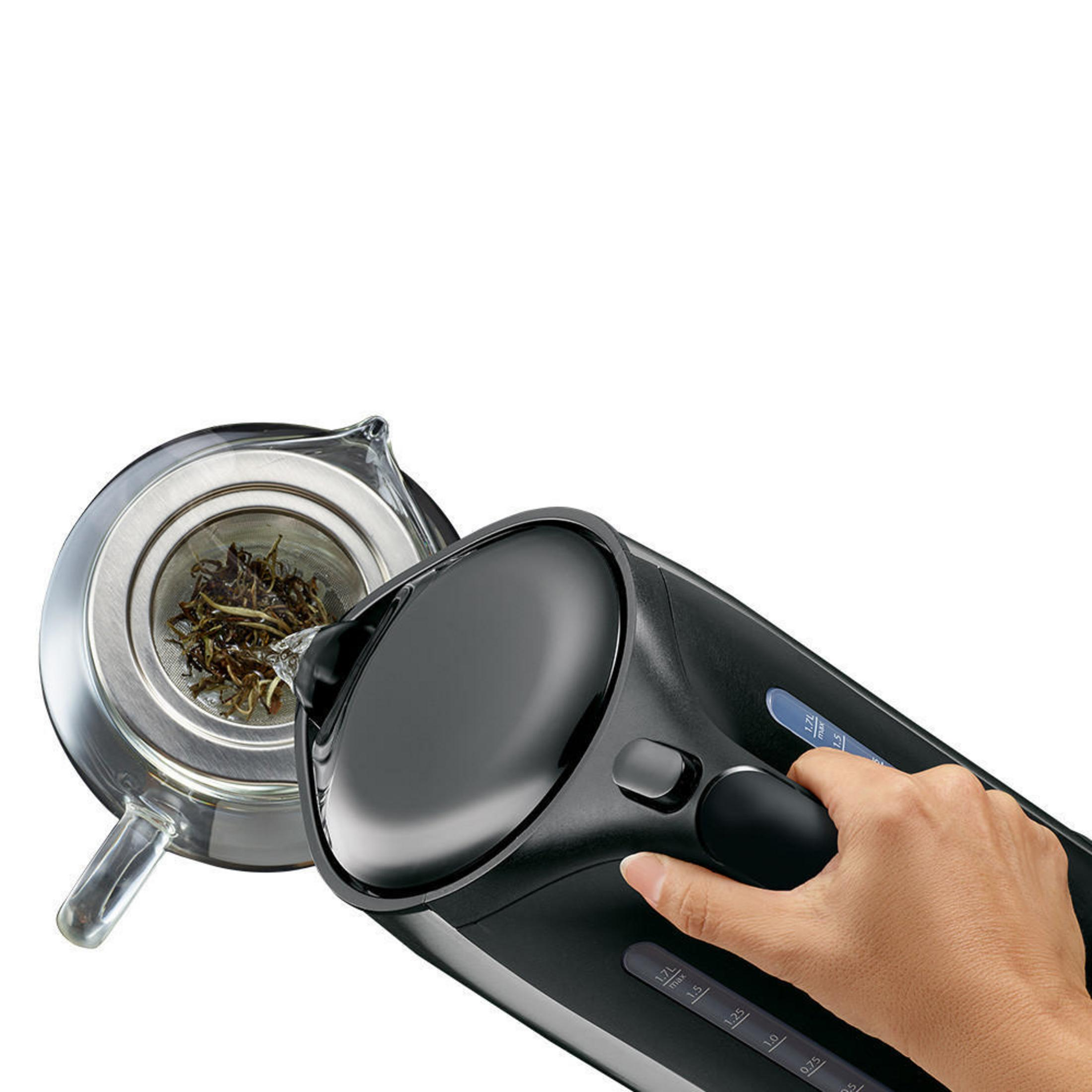 Bollitore elettrico nero che versa acqua in un infusore per tè in vetro. Una mano tiene il bollitore.