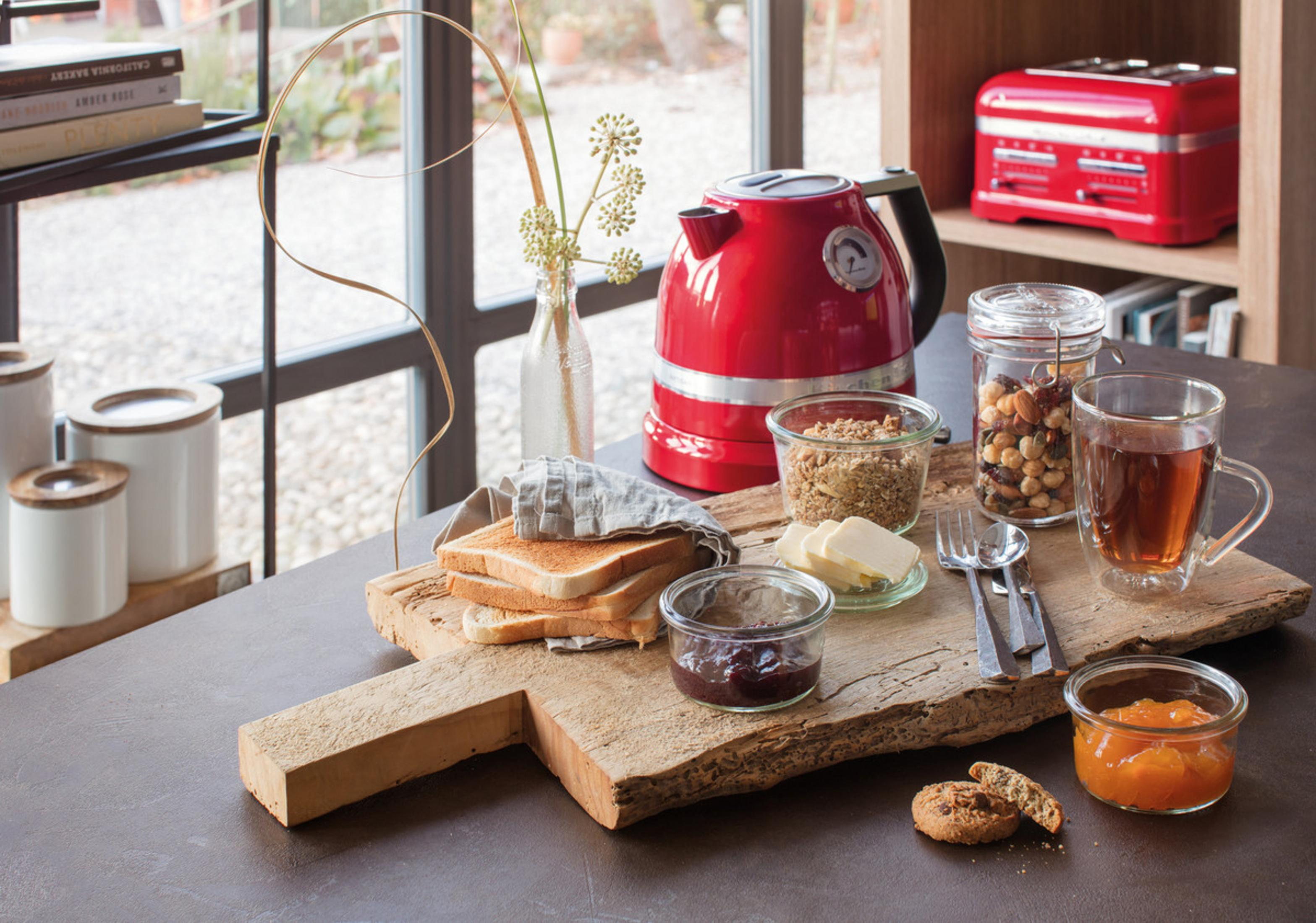Stół śniadaniowy z czerwonym czajnikiem, tostami, dżemem, orzechami i herbatą.
