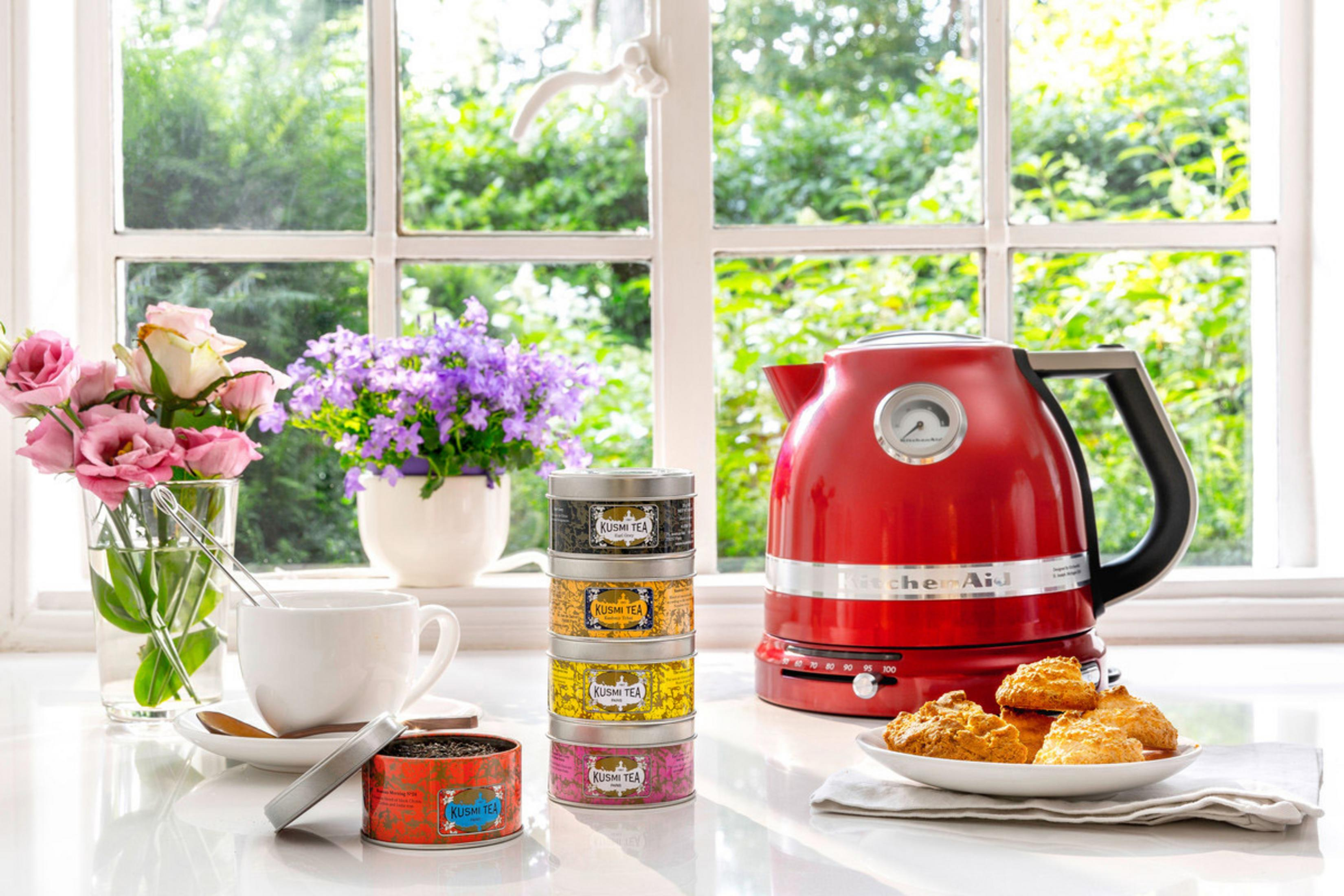 Scena kuchenna z czerwonym czajnikiem, puszkami herbaty, kwiatami i ciasteczkami na parapecie.