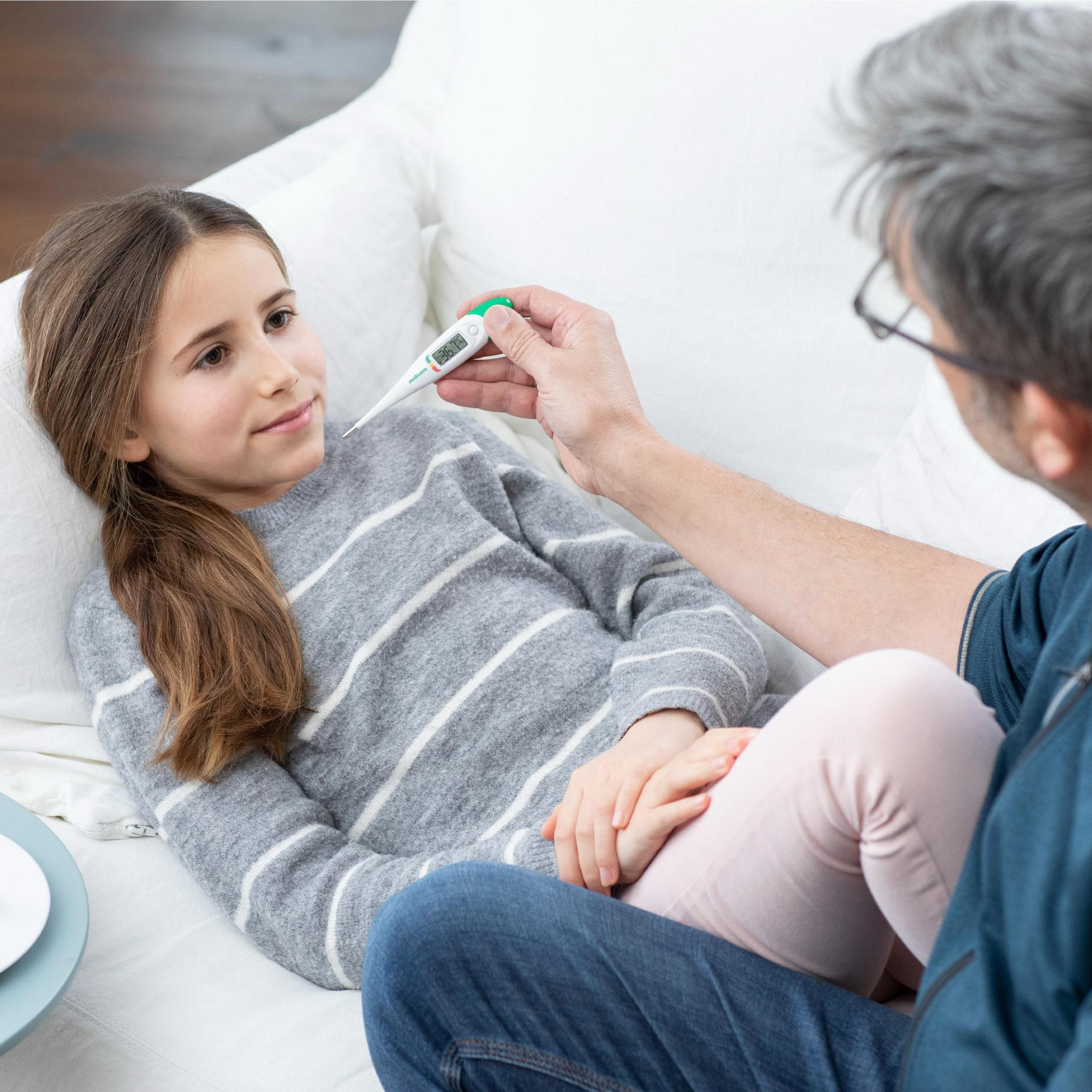 Dziewczynce leżącej na białej kanapie mężczyzna mierzy temperaturę termometrem cyfrowym.