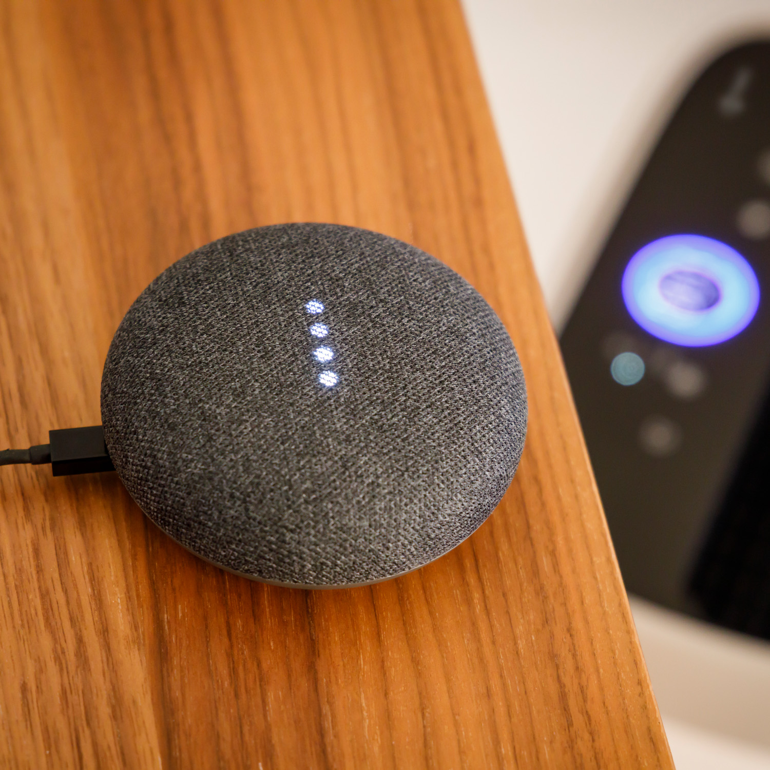 Un altavoz inteligente gris con luces blancas sobre una mesa de madera. Un mando a distancia en el fondo.