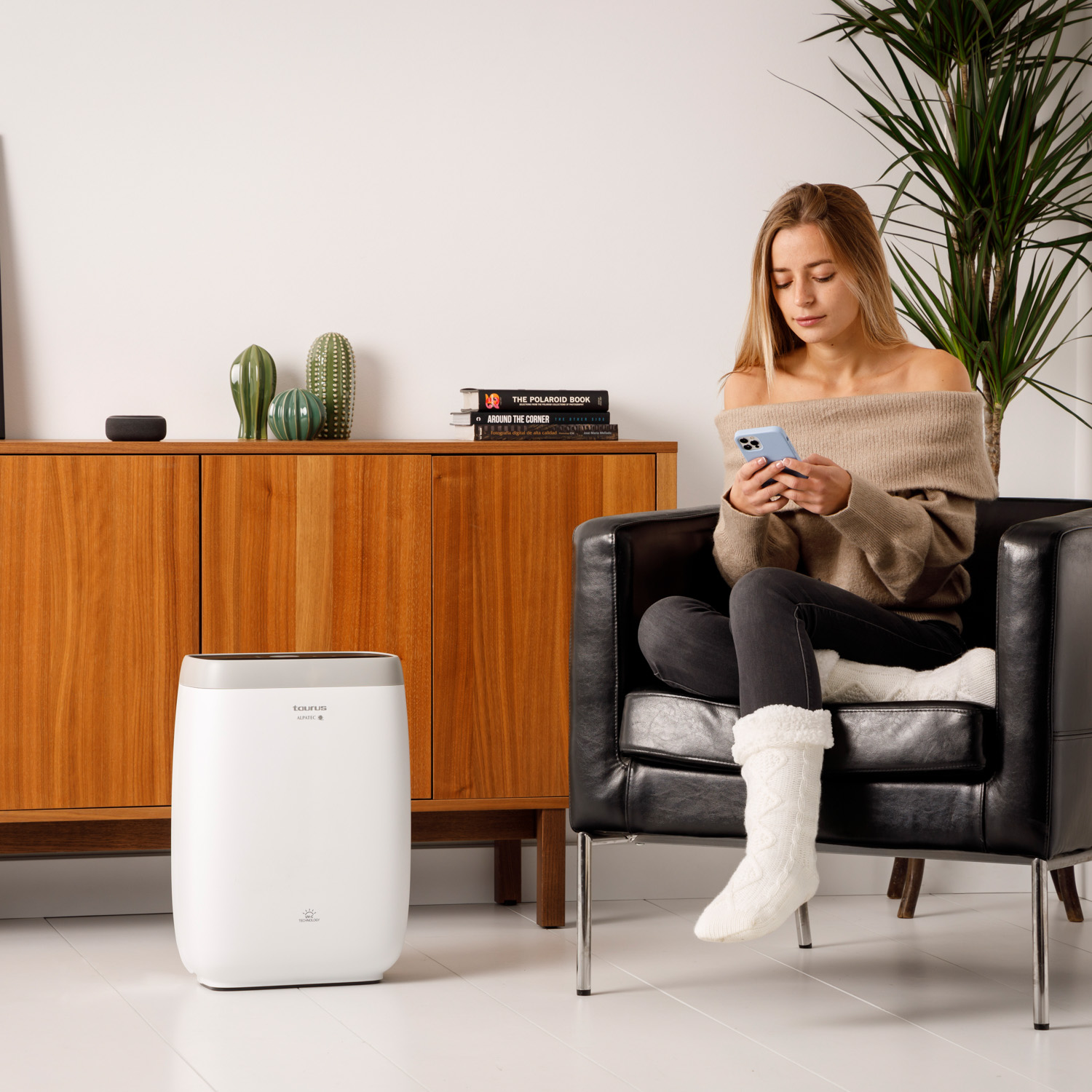 Mujer sentada en una silla, usando su teléfono. Purificador de aire y plantas al fondo.