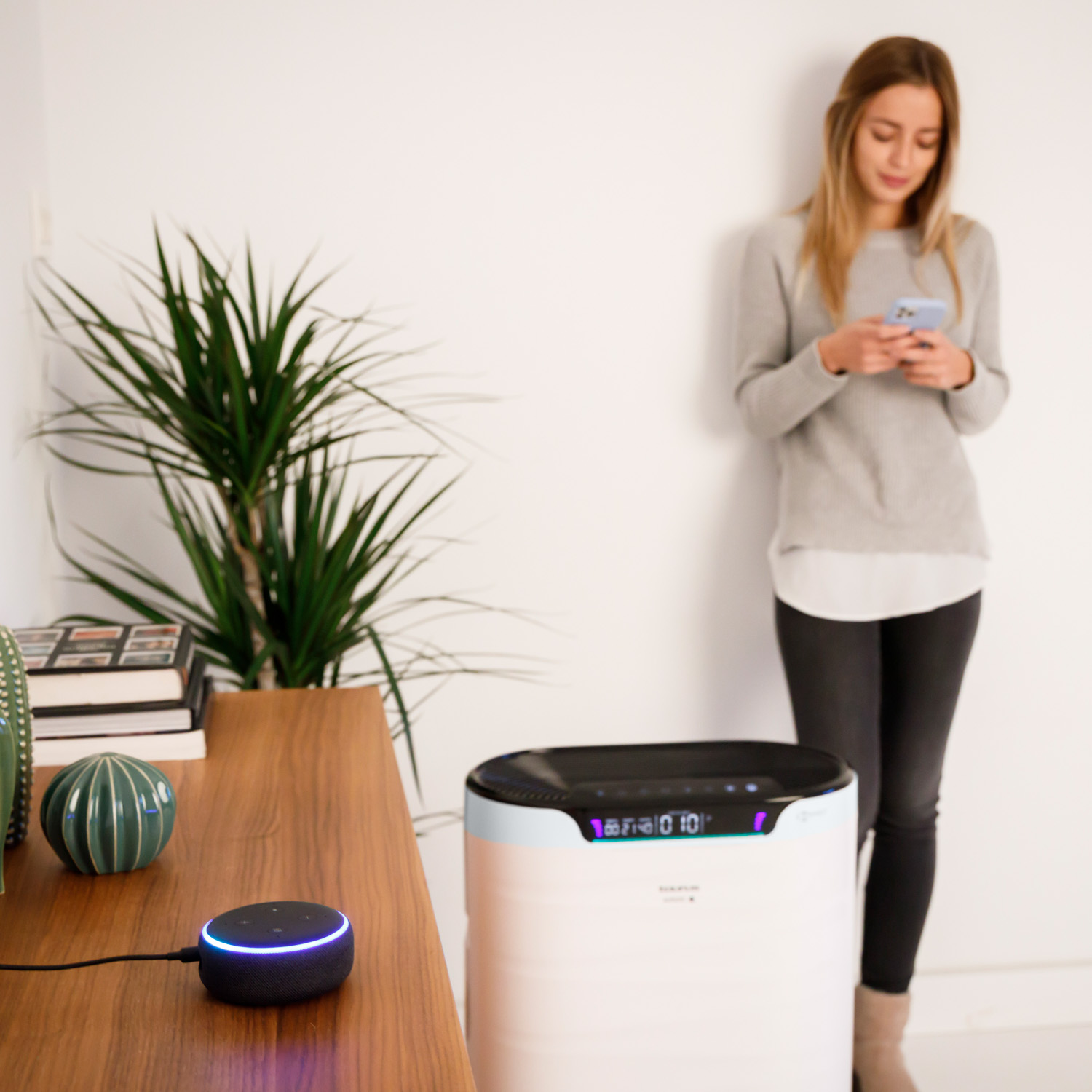 Una mujer está cerca de un purificador de aire usando su teléfono; un altavoz inteligente es visible.