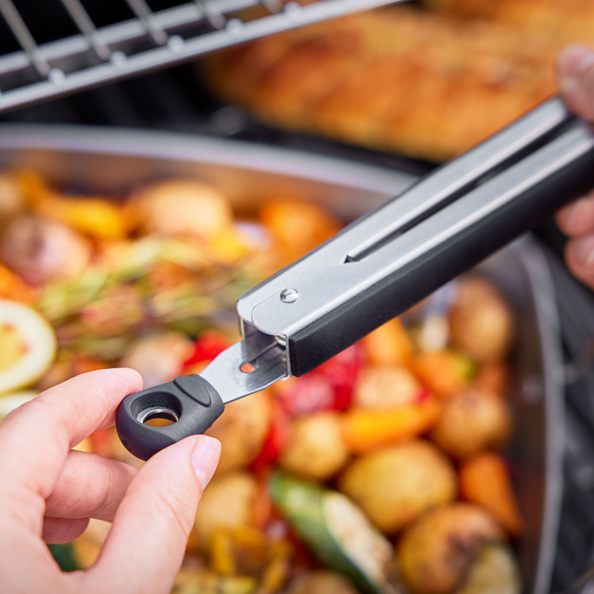 Eine Hand hält eine Zange über einem Tablett mit gegrilltem Gemüse, unscharfer Hintergrund.
