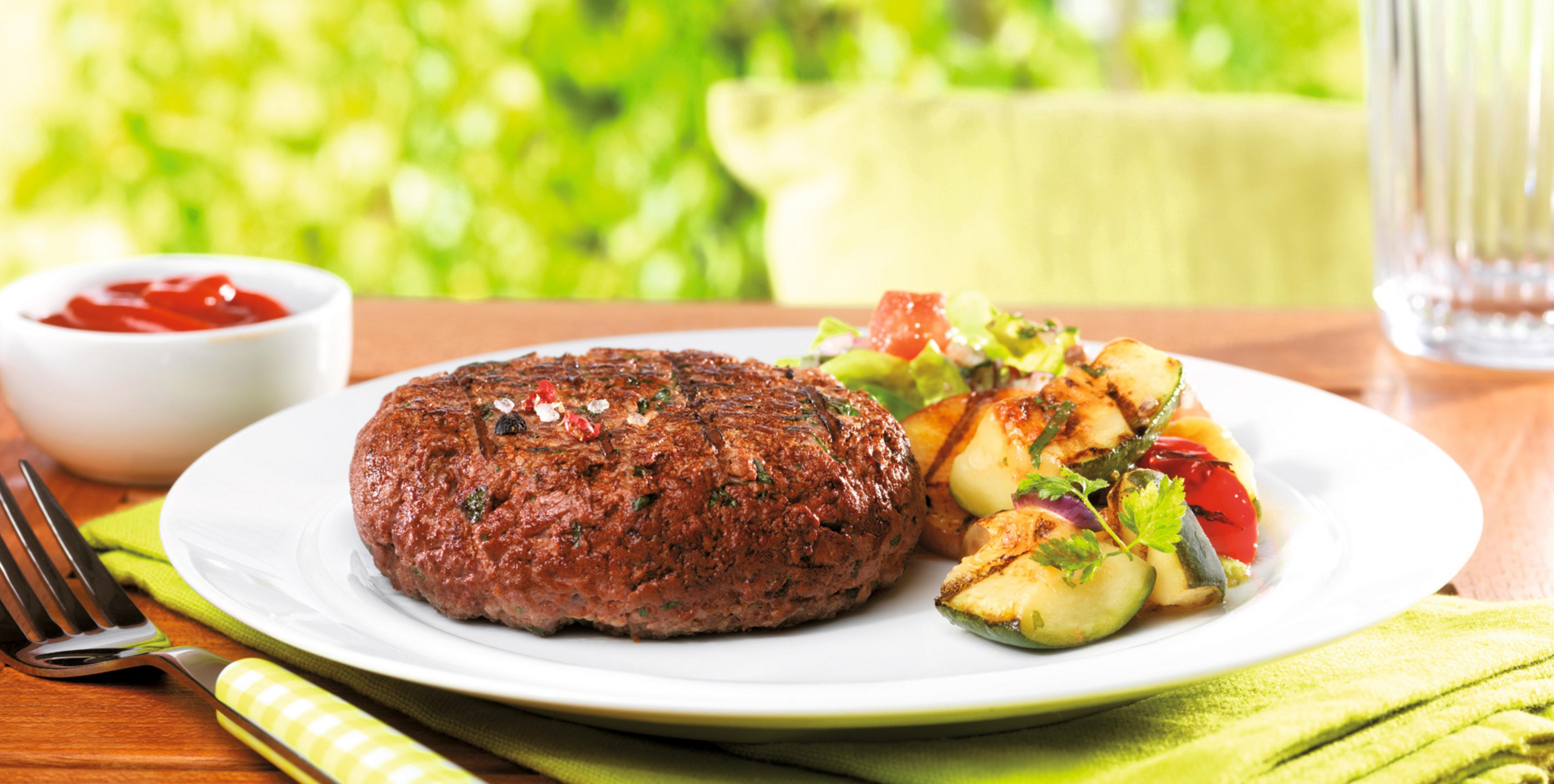 Teller mit Burger und gegrilltem Gemüse. Ketchup und verschwommener grüner Hintergrund.