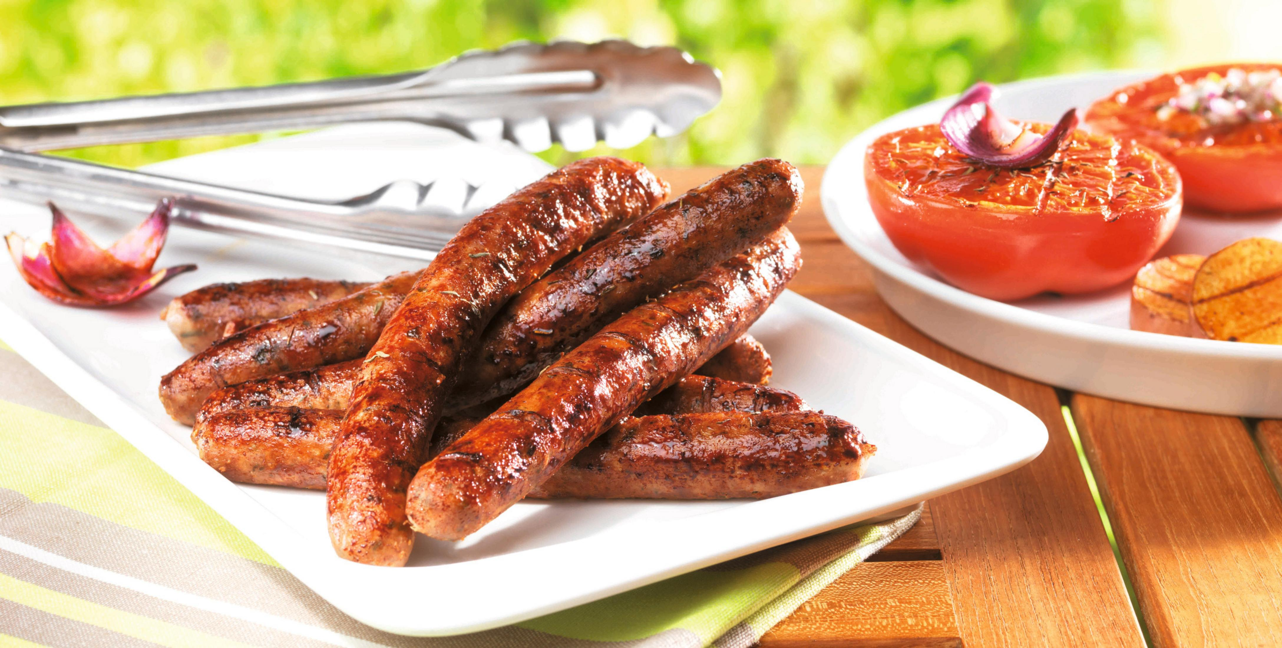 Gegrillte Würstchen und Tomaten auf weißem Teller auf Holztisch, Zange daneben.