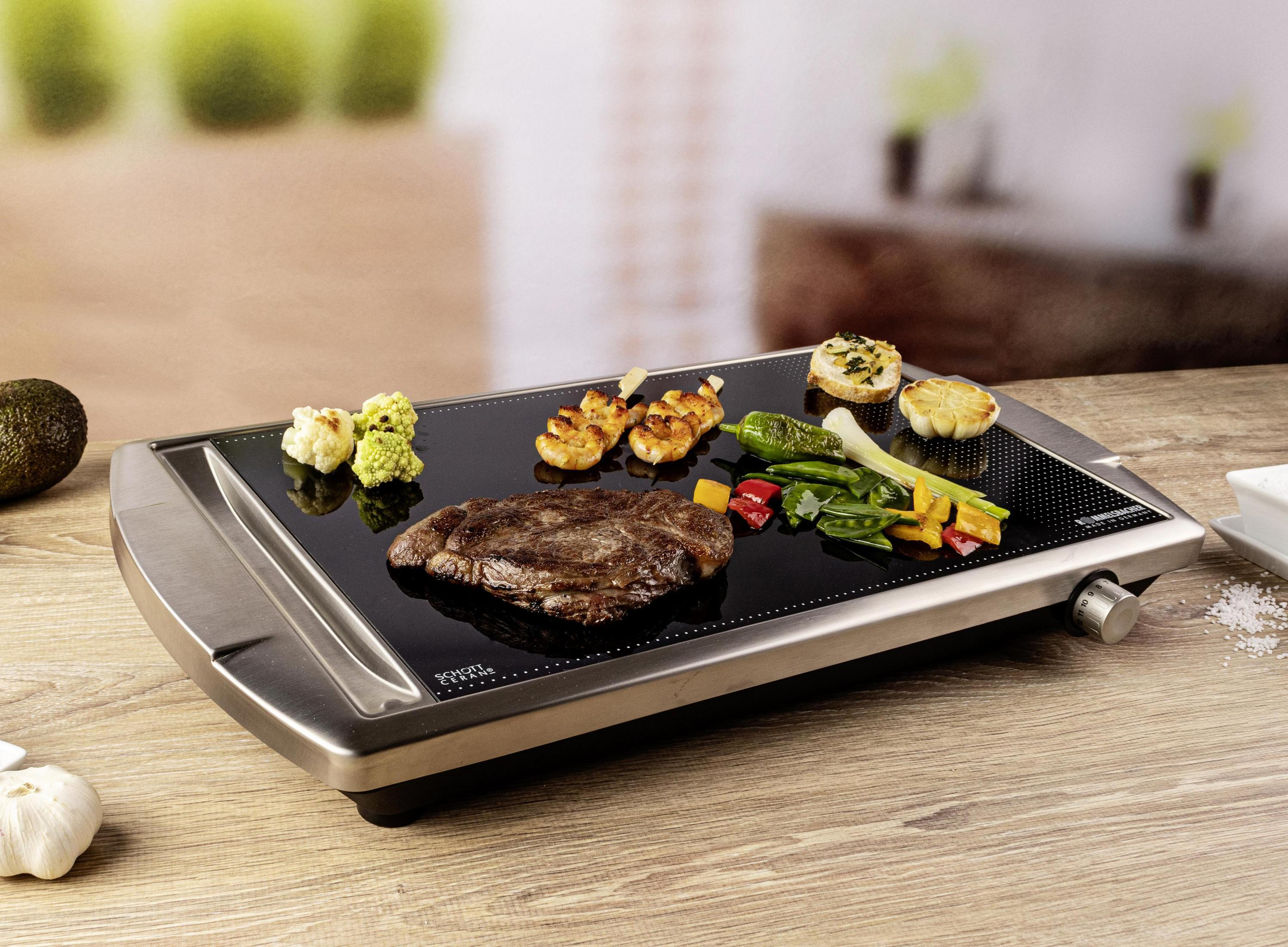 Elektrische Grillplatte mit Steak, Gemüse und Avocado auf schwarzer Oberfläche, auf einem Holztisch.