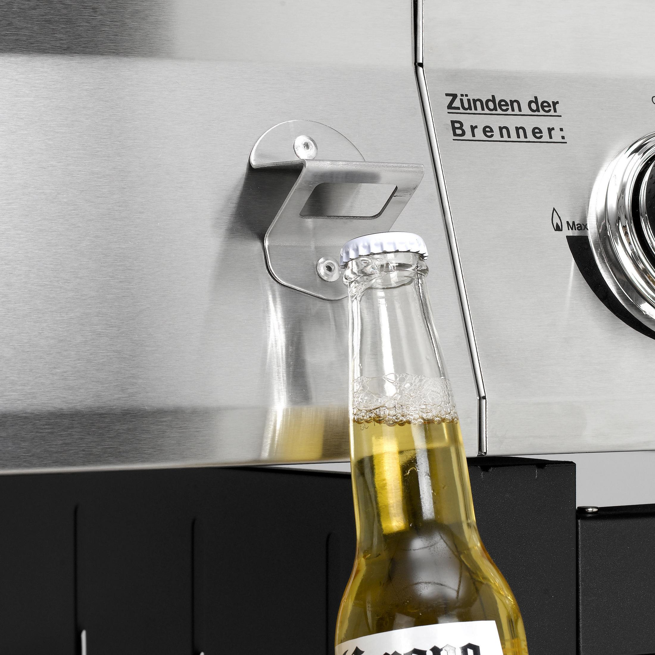 Nahaufnahme einer Bierflasche, die mit einem Flaschenöffner am Grill geöffnet wird.