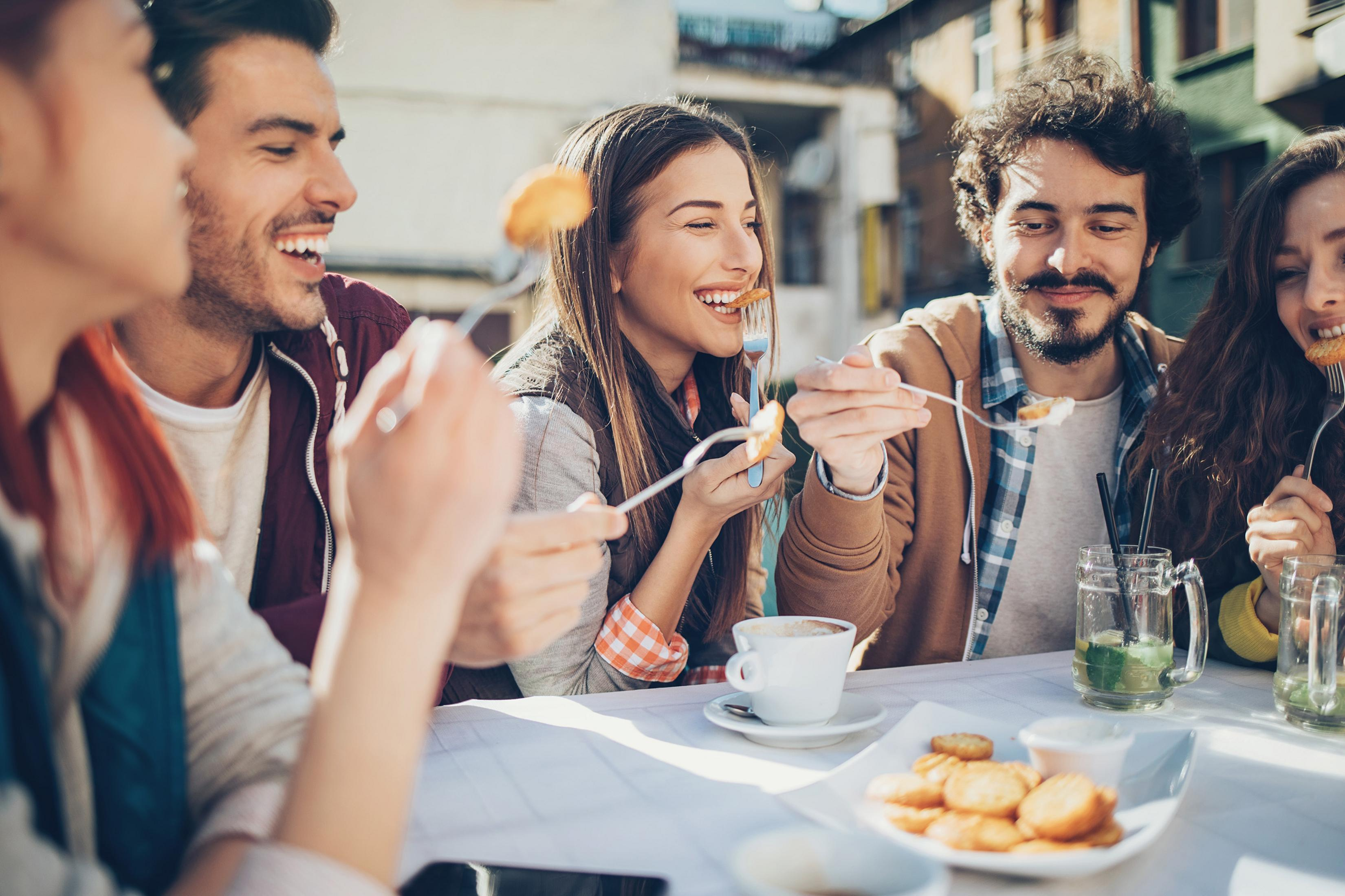 Freunde essen draußen, lächeln. Tisch mit Essen und Getränken im Fokus.