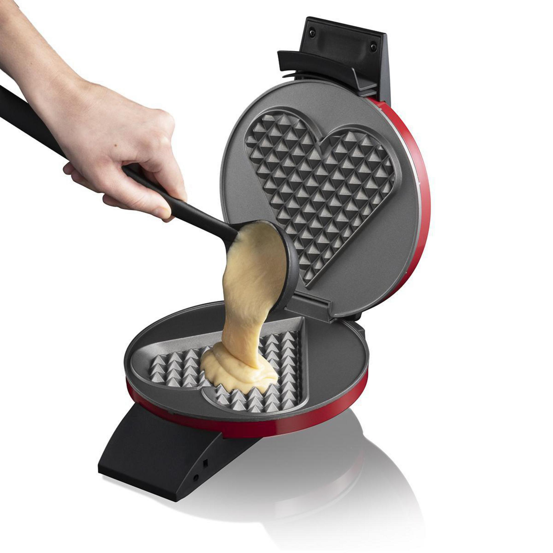 Ręka wlewa ciasto do gofrownicy w kształcie serca. Gofrownica jest czerwona i otwarta.