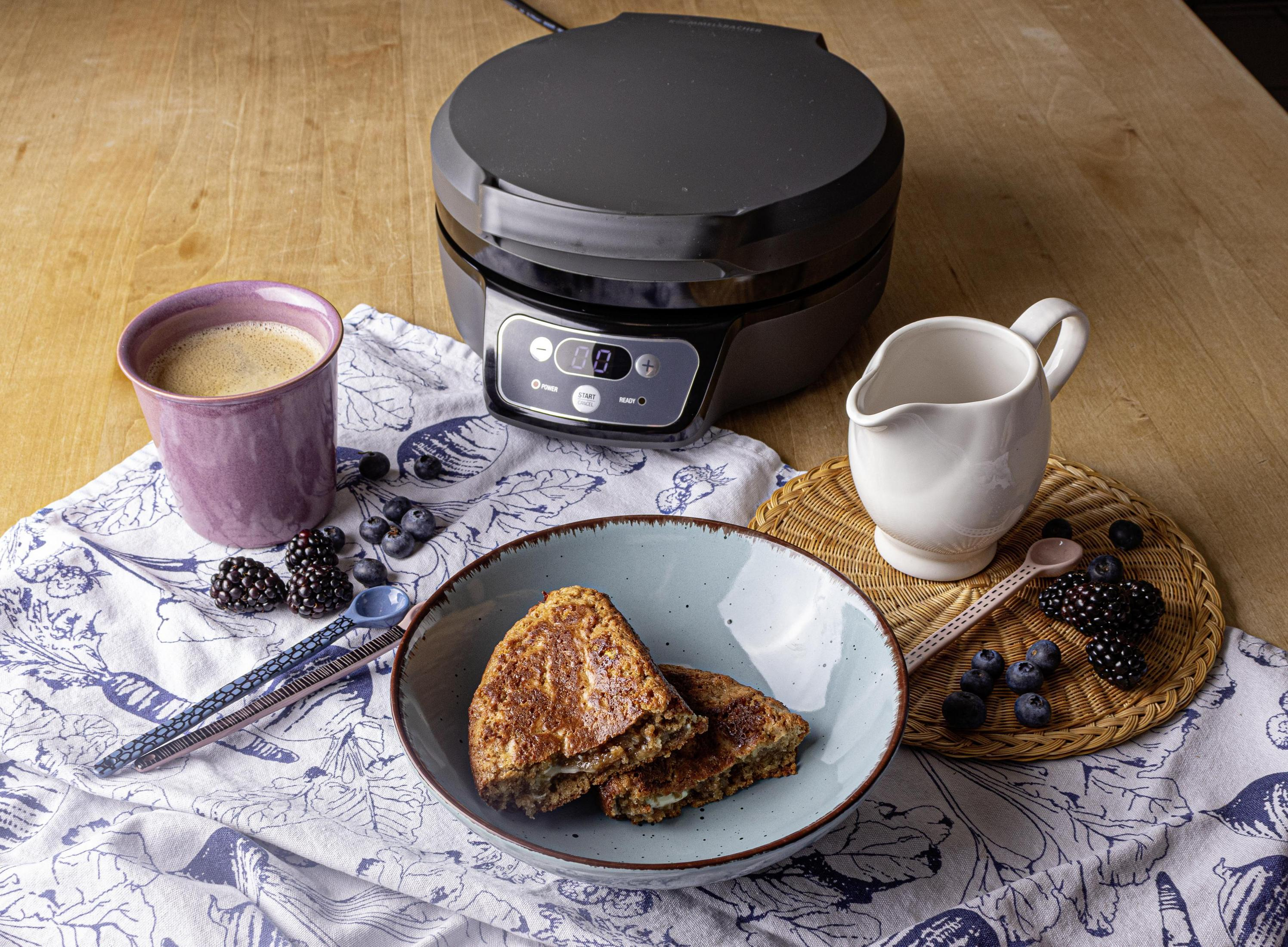 Ein Waffeleisen mit Waffeln, eine Kaffeetasse und Beeren auf einem Holztisch.