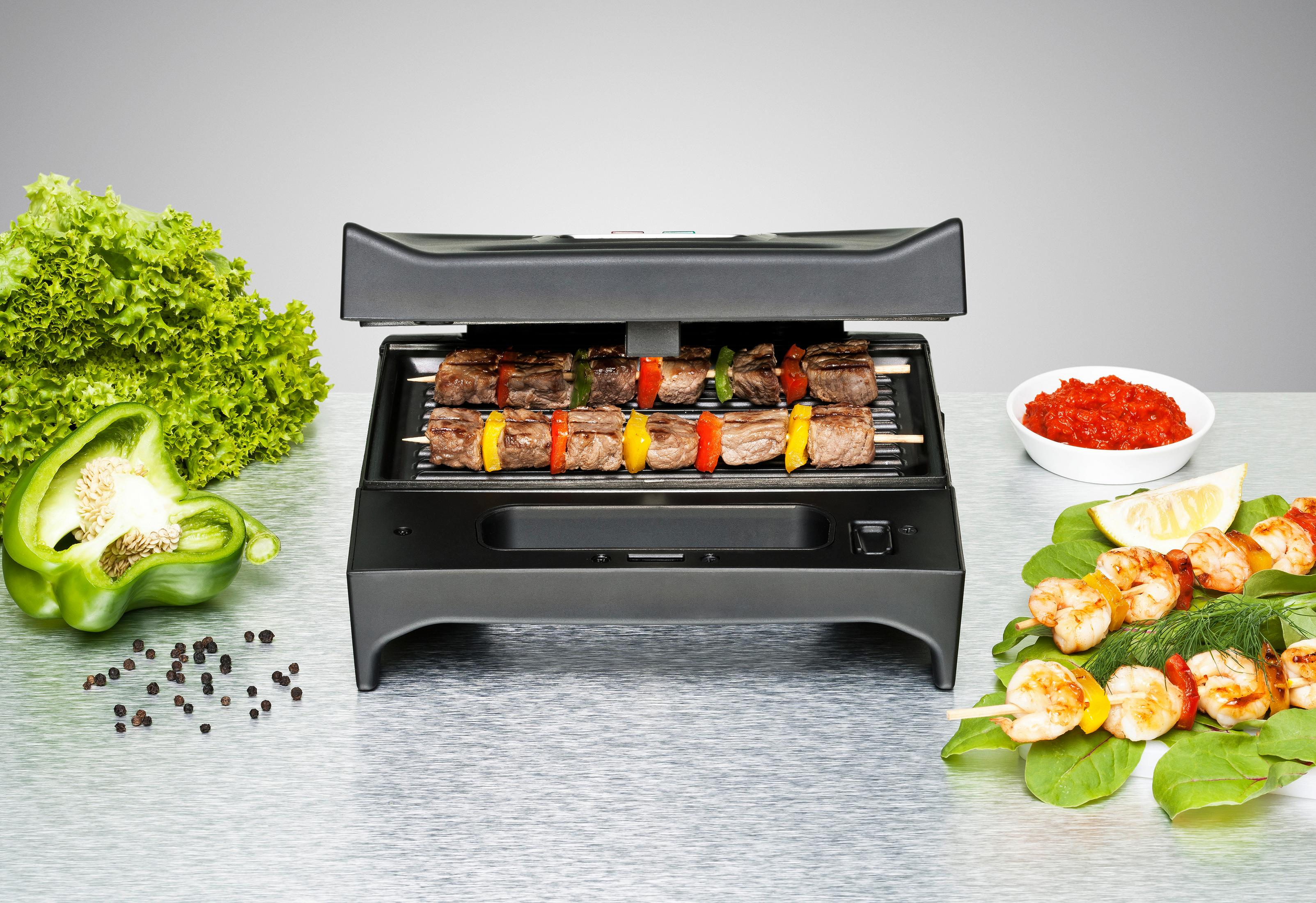 Una parrilla eléctrica con brochetas de carne y verduras, rodeada de verduras y camarones.