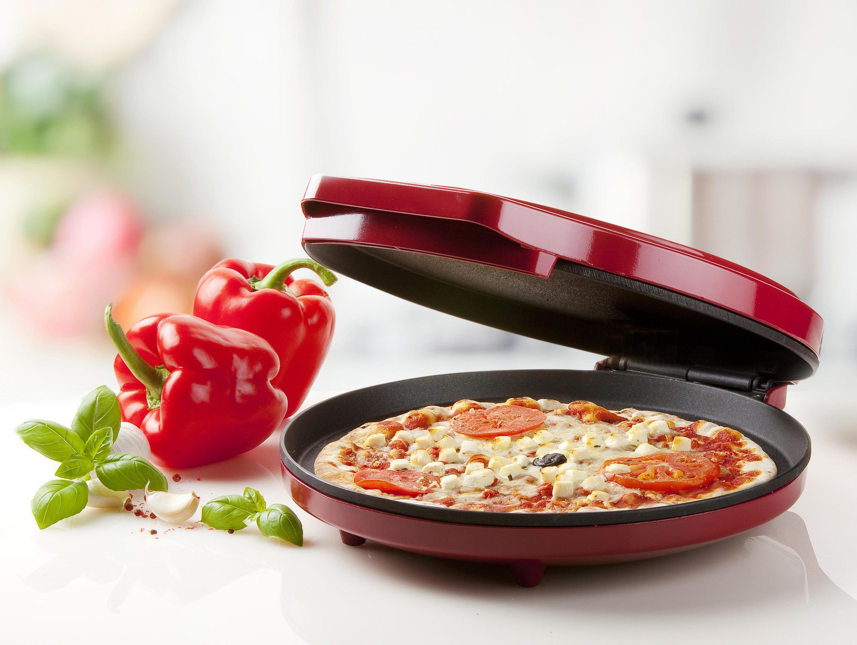 Una máquina de pizza roja con una pizza dentro. Pimientos rojos y hojas de albahaca están al lado.