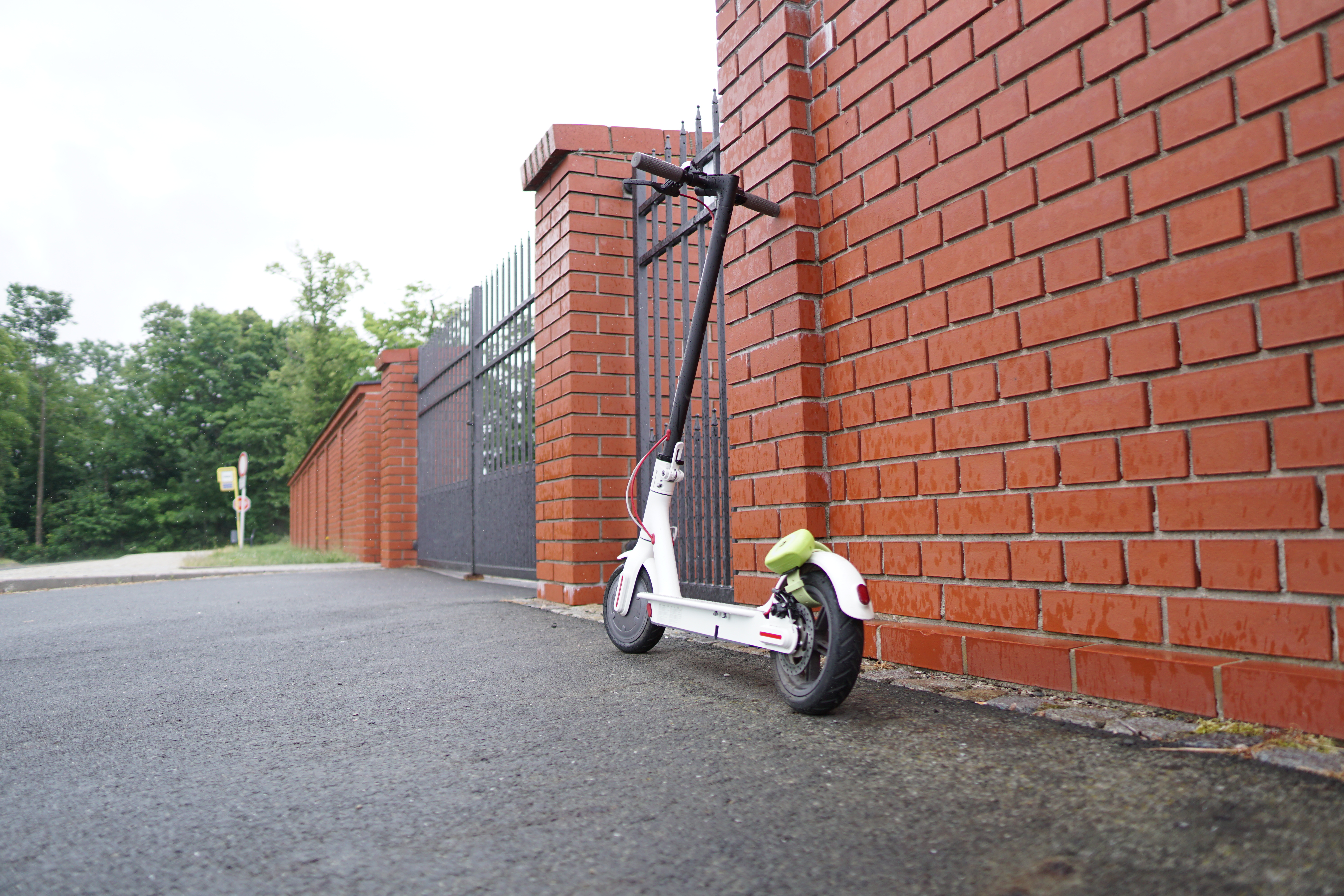 Ein weißer Roller lehnt an einer Backsteinmauer neben einem Metallzaun und einer Straße.