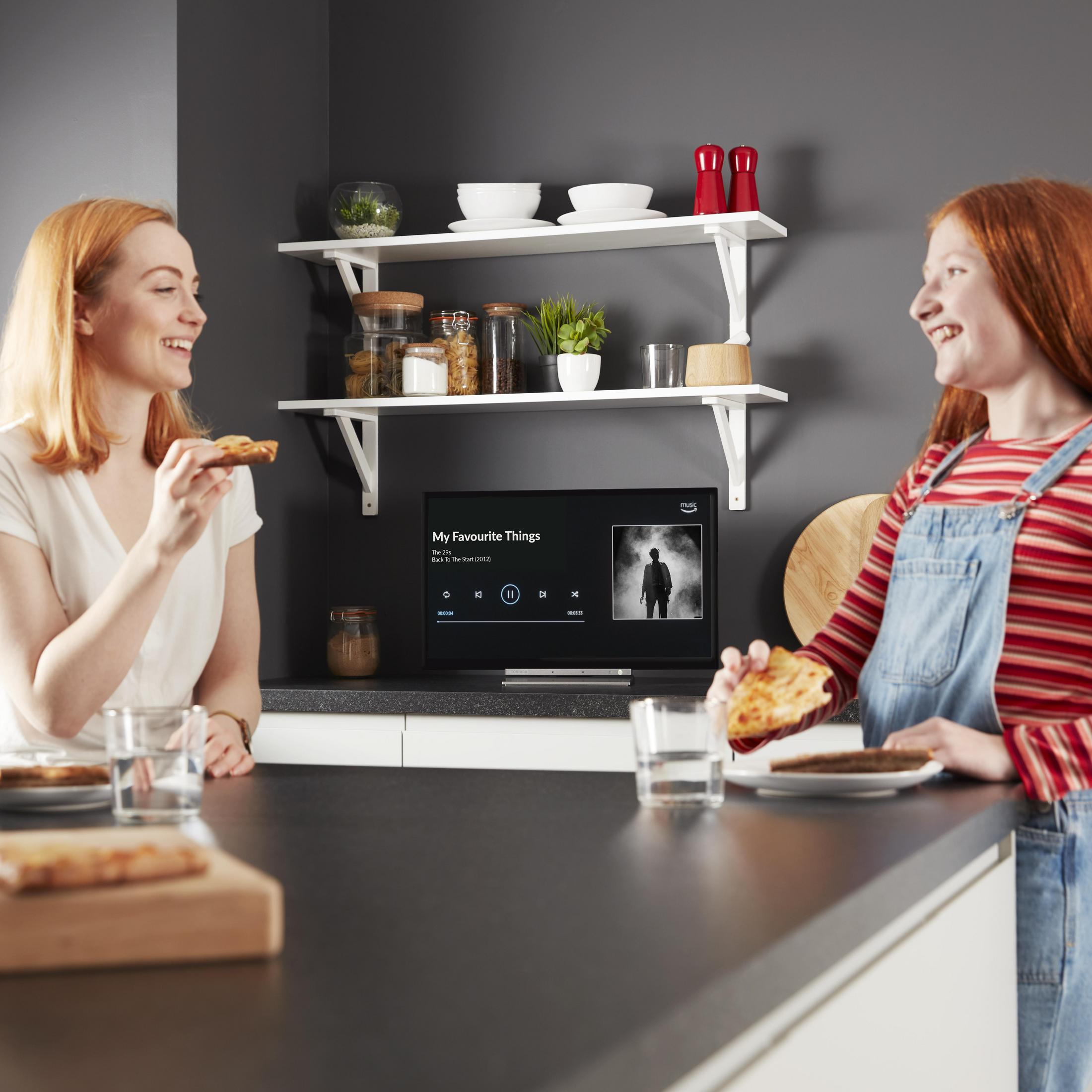 Due donne in cucina guardano la TV, mangiando pizza.