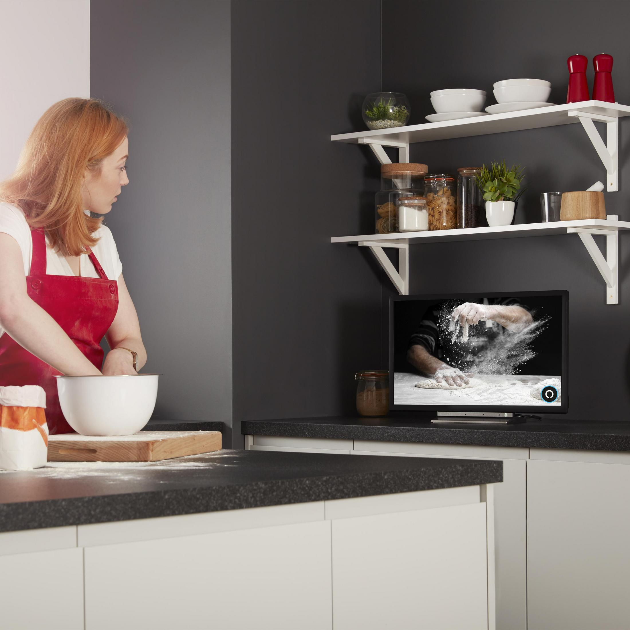 Una donna in cucina, che guarda un monitor che mostra una scena di cucina, preparando l'impasto.