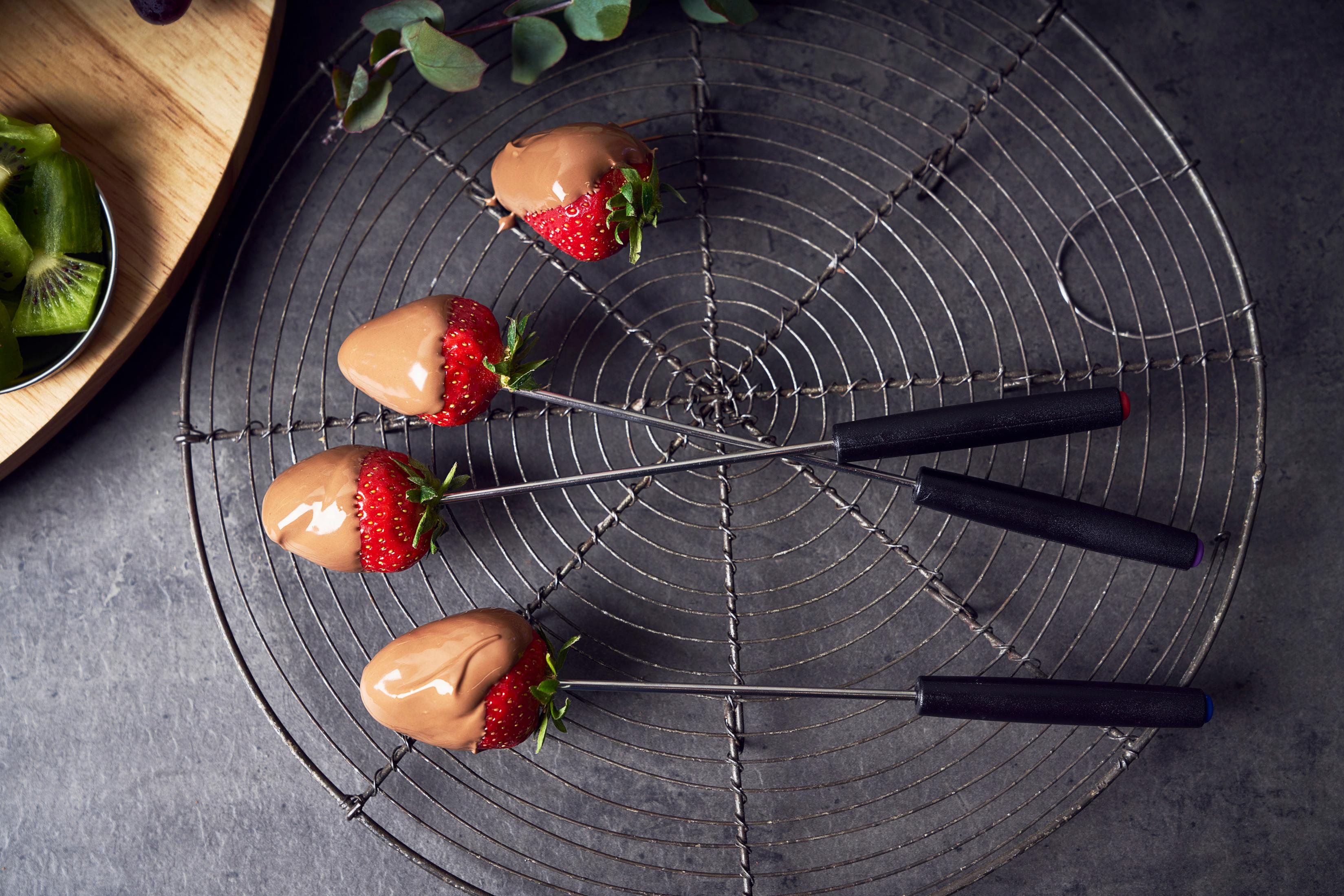 Tre fragole immerse nel cioccolato su una griglia metallica con forchette per intingere.