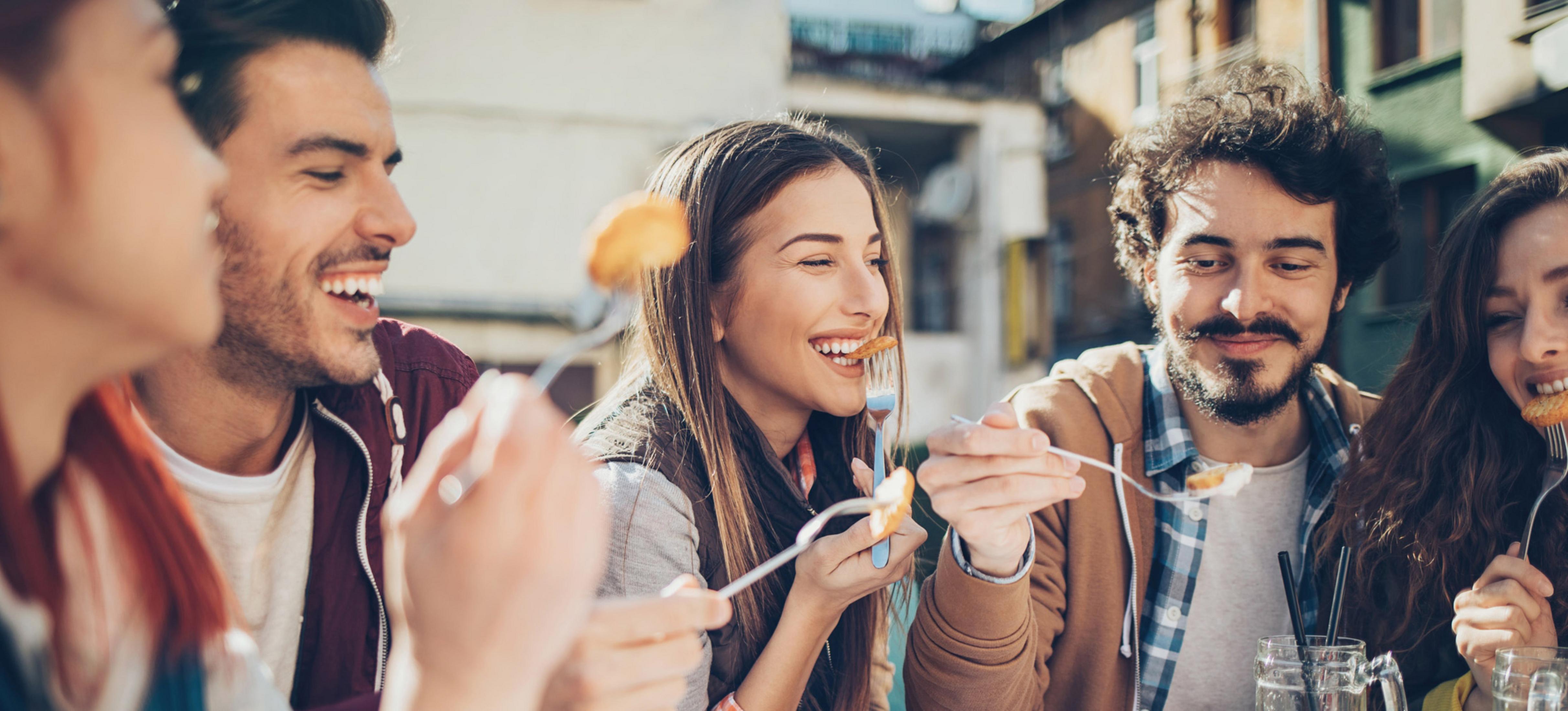 Gruppo di persone che ridono mentre mangiano all'aperto, giornata di sole.