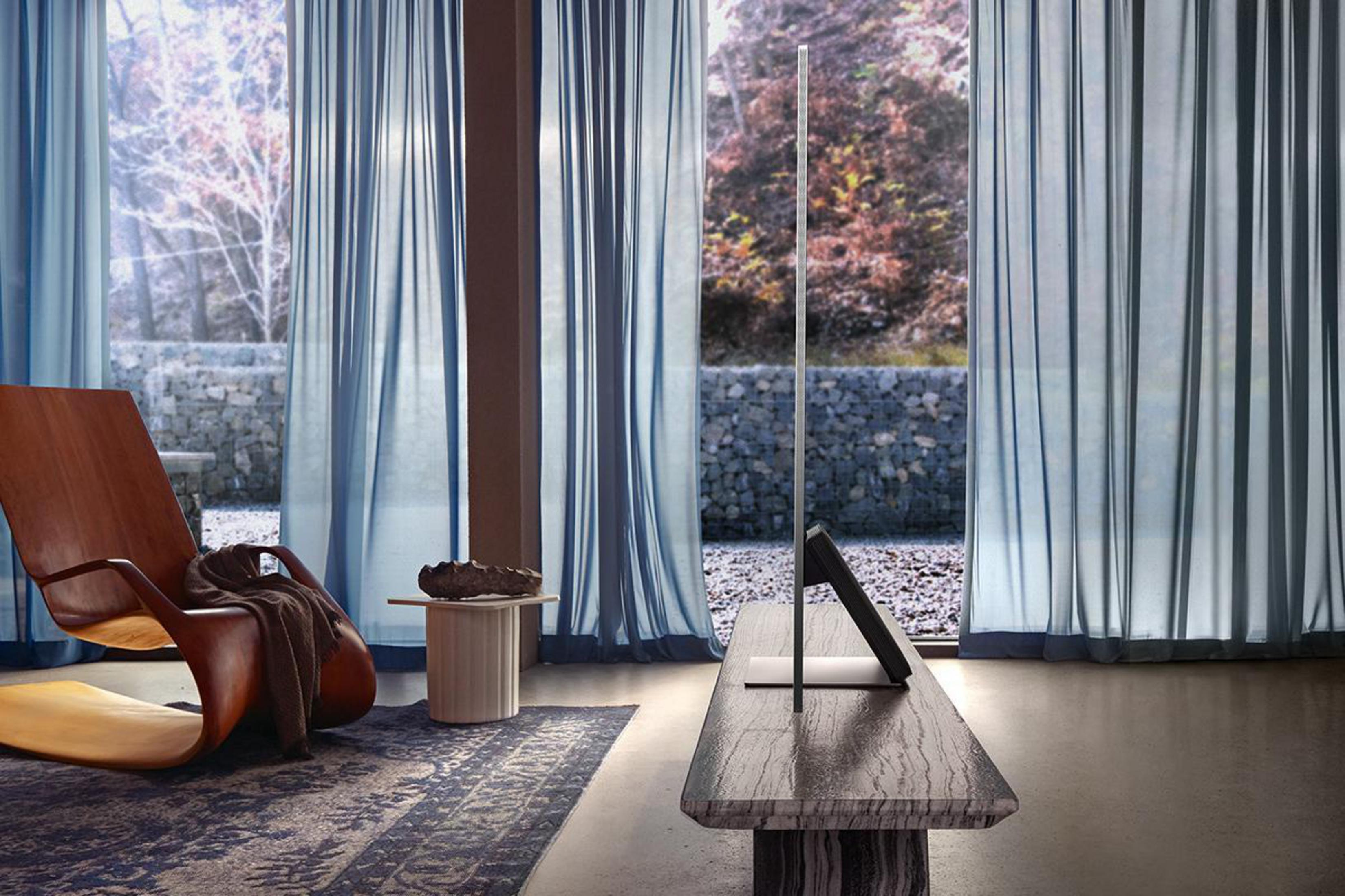 Nowoczesny telewizor stoi pionowo, z jasnym tłem i zasłonami, w pobliżu drewnianego krzesła i stołu.