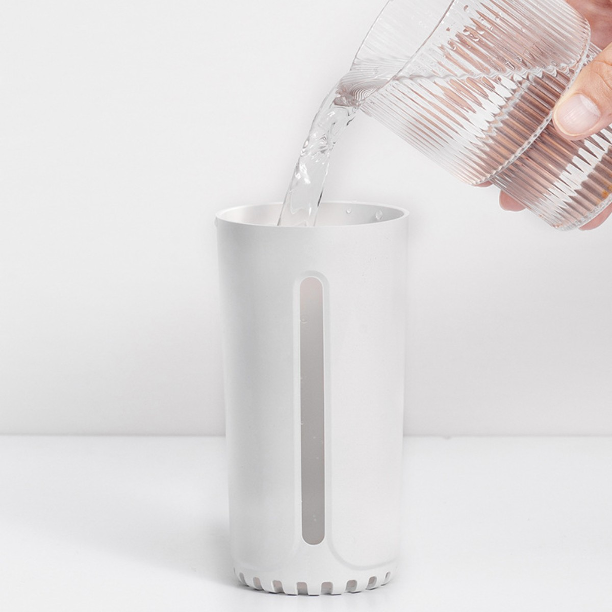 Agua vertiéndose en un recipiente cilíndrico blanco. Una mano sostiene un vaso transparente.