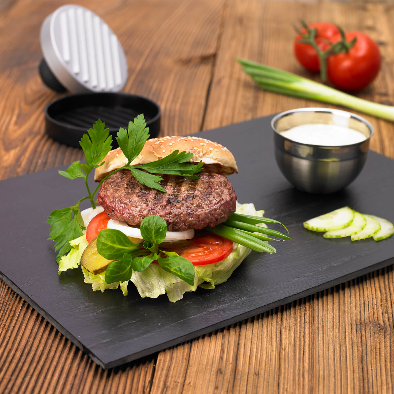 Ein Burger auf schwarzem Schiefer, mit Tomaten, Salat und weißer Soße, auf einem Holztisch.