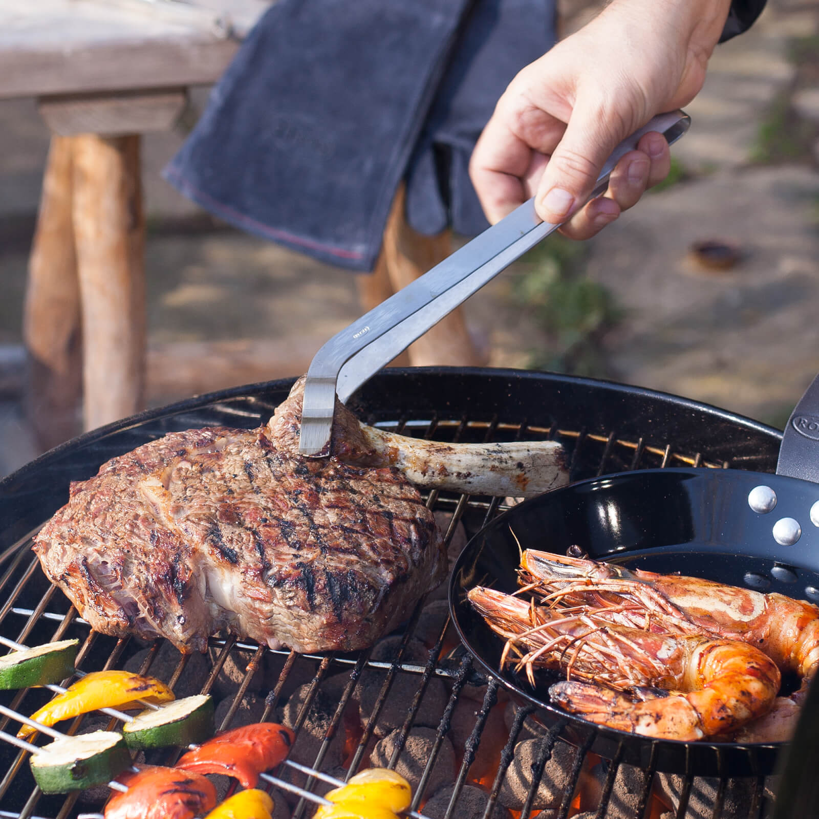Eine Hand hält eine Zange über einem Grill mit Steak und Garnelen. Gemüse wird auf dem Rost gegrillt.