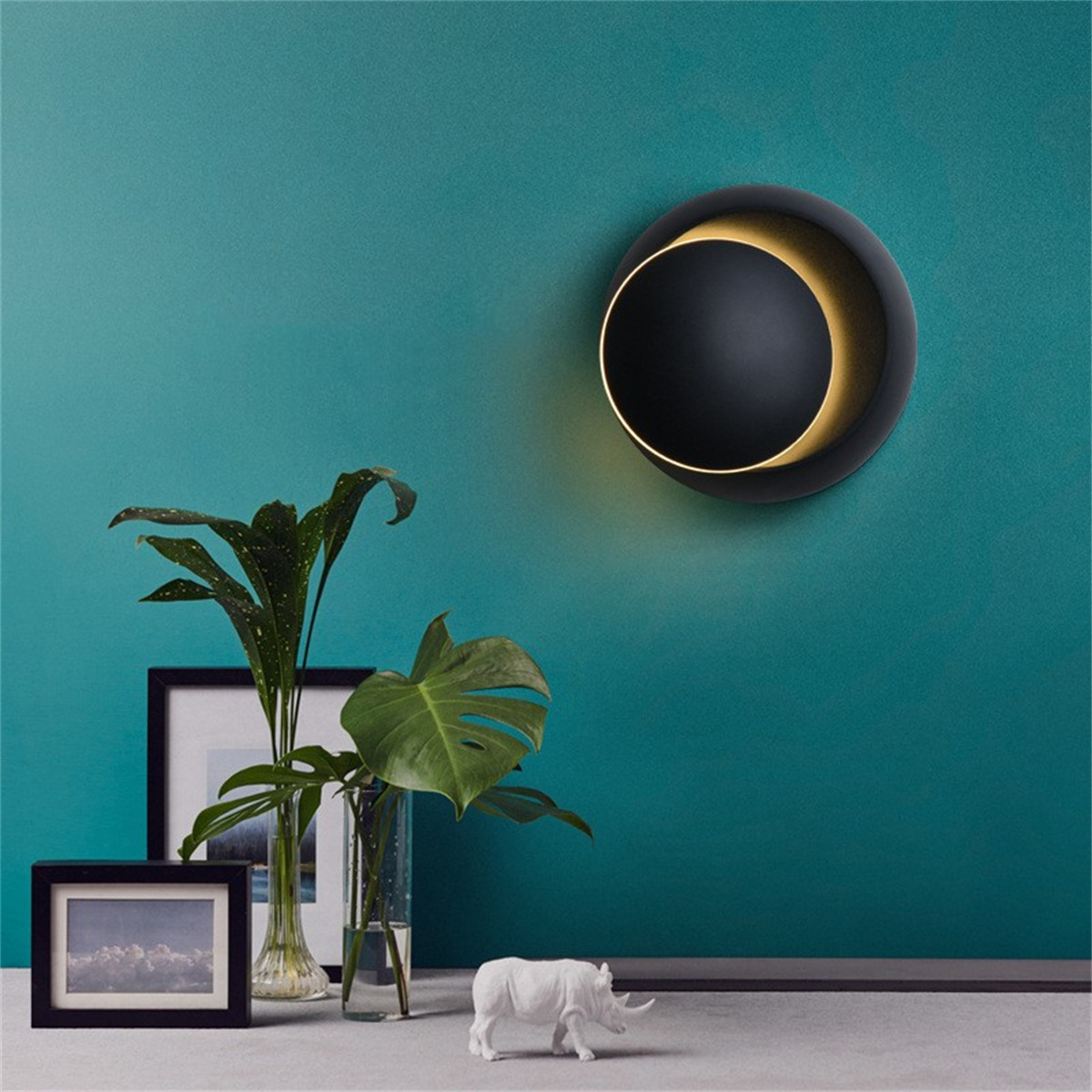 Eine Wand mit einer schwarzen, halbmondförmigen Lampe. Ein Bild, eine Pflanze und eine kleine Nashornstatue.