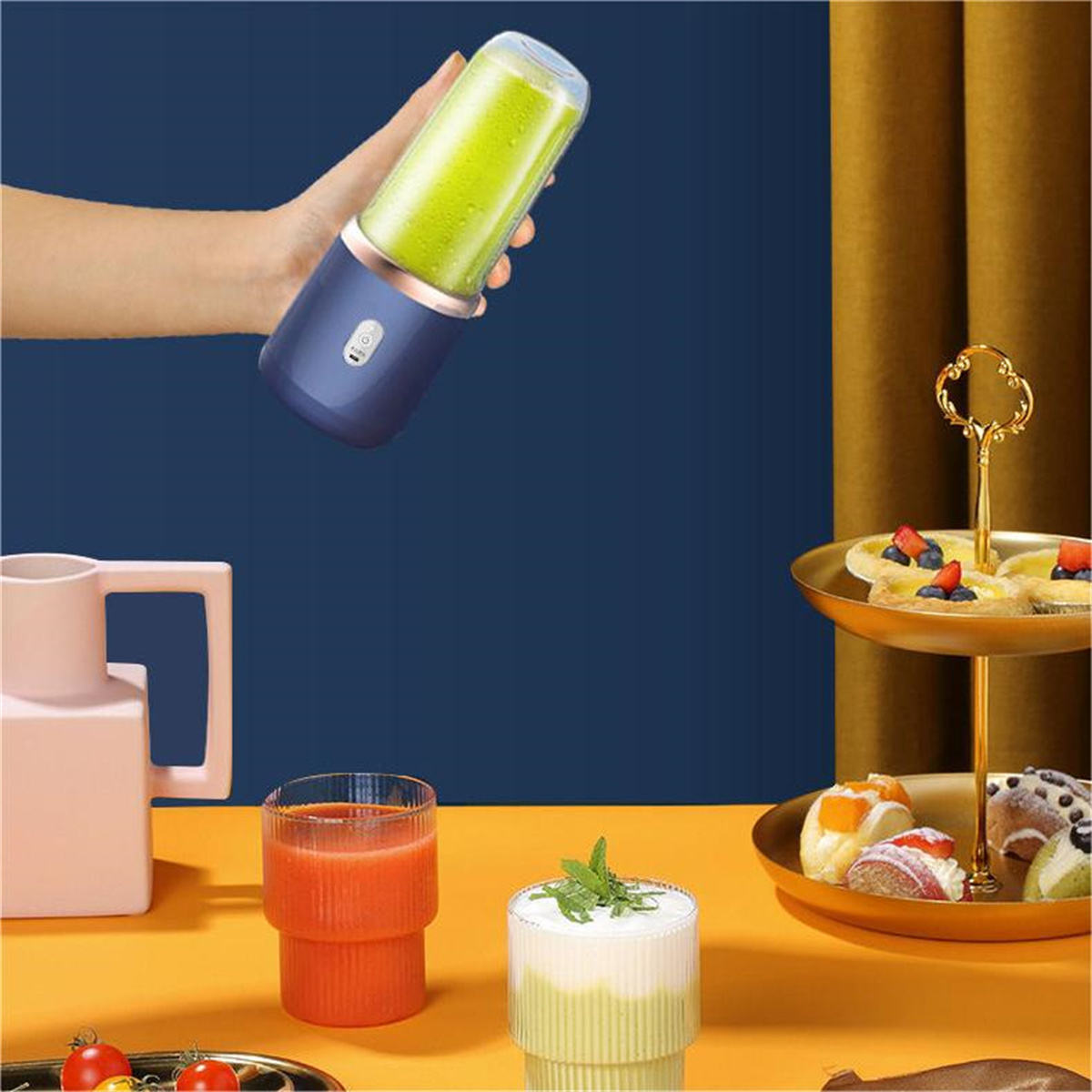 Hand hält Mixer mit grüner Flüssigkeit, mit Saft und Desserts auf einem Tisch.