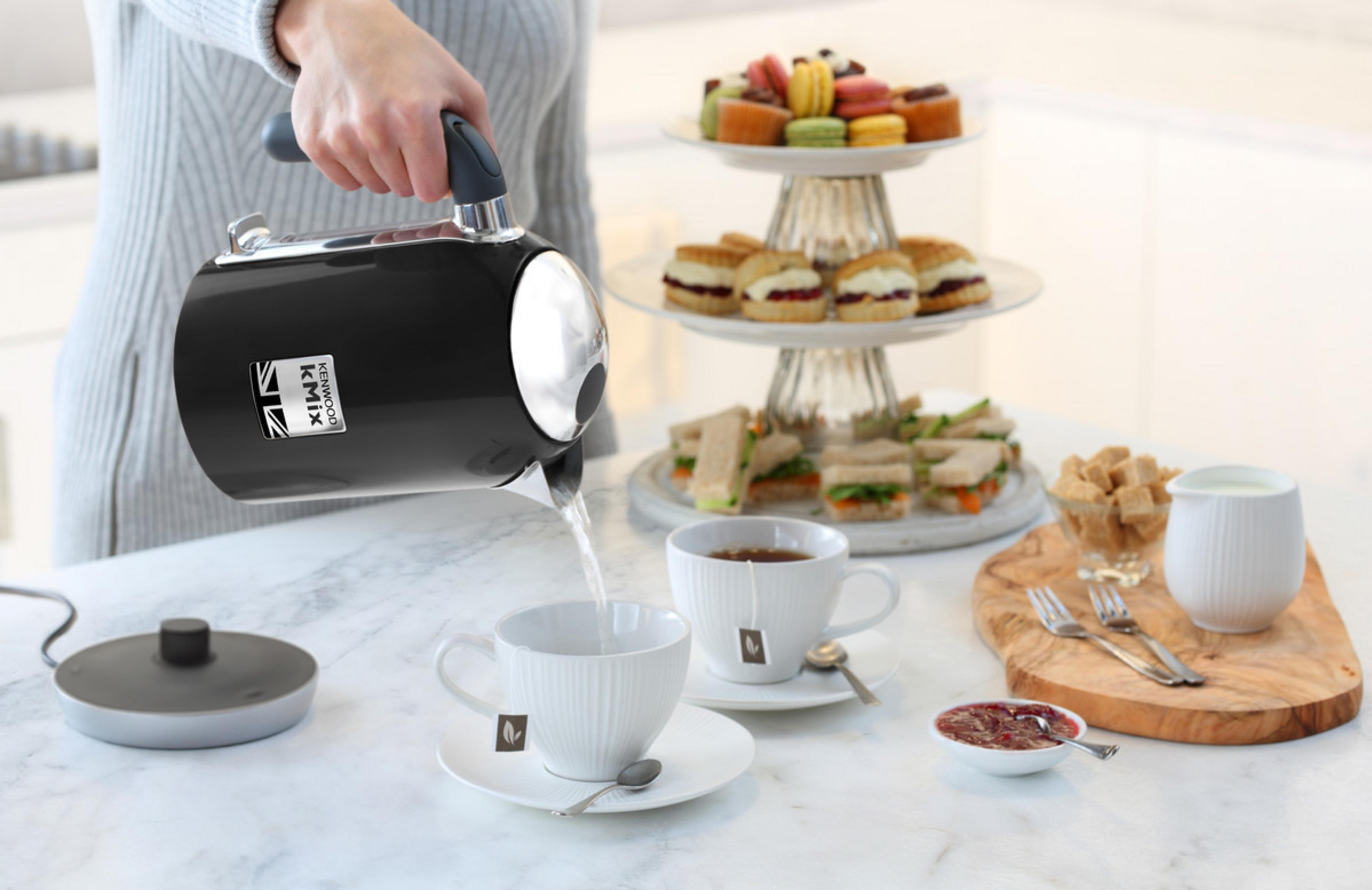 Kobieta nalewa wodę z czarnego czajnika do filiżanki herbaty. Kanapki i ciasta w tle.