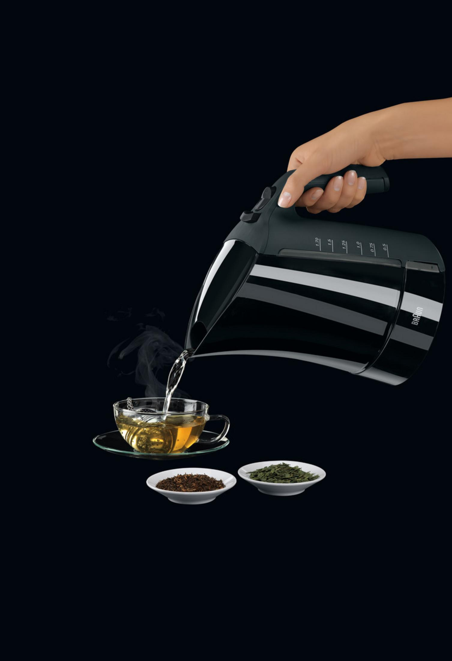 Eine Hand gießt Wasser aus einem schwarzen Wasserkocher in eine Glastasse mit Tee. Zwei Schalen mit Teeblättern daneben.