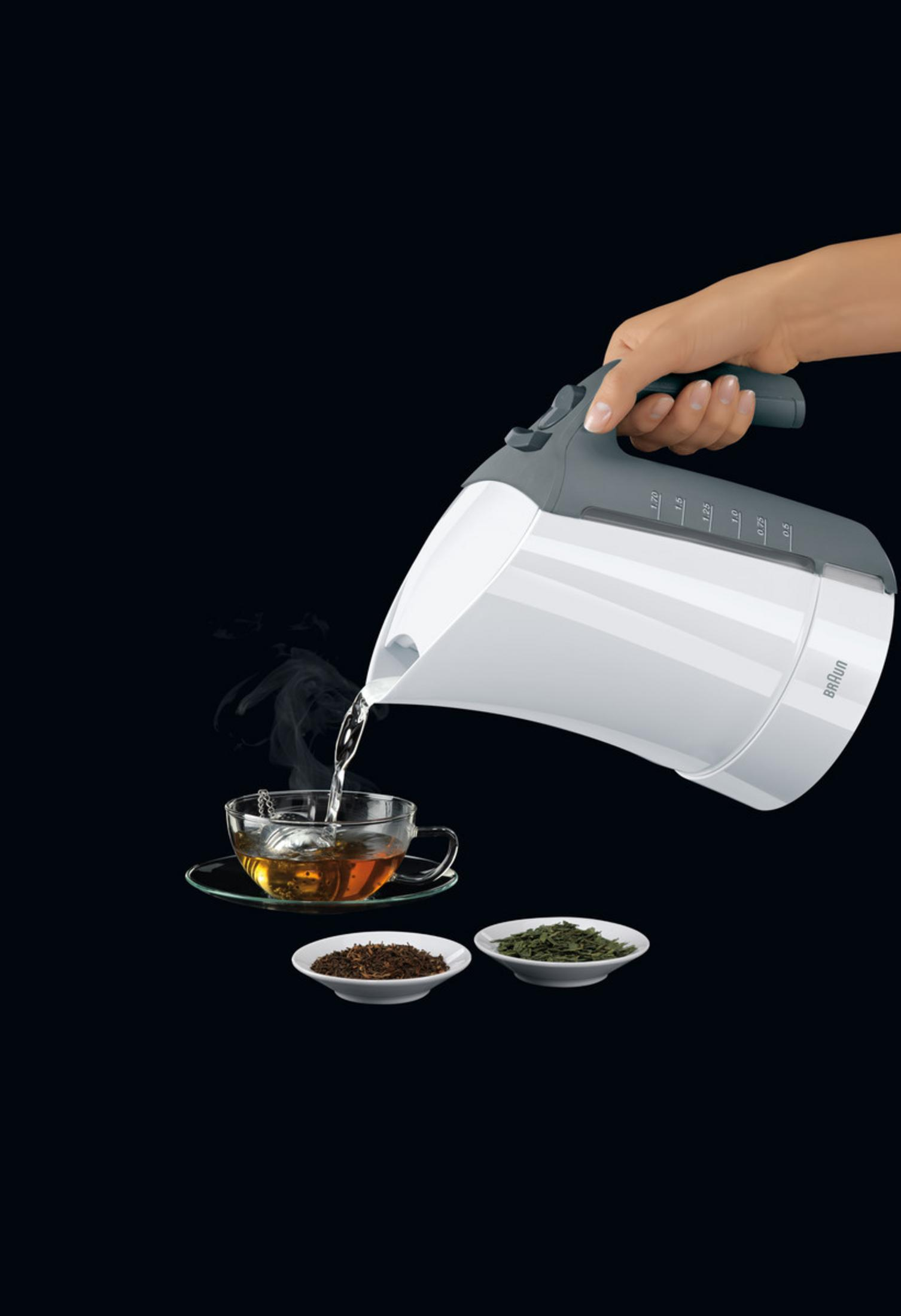 Ein weißer Wasserkocher gießt Wasser in eine Teetasse mit Teeblättern. Daneben zwei kleine Schalen mit Teeblättern.