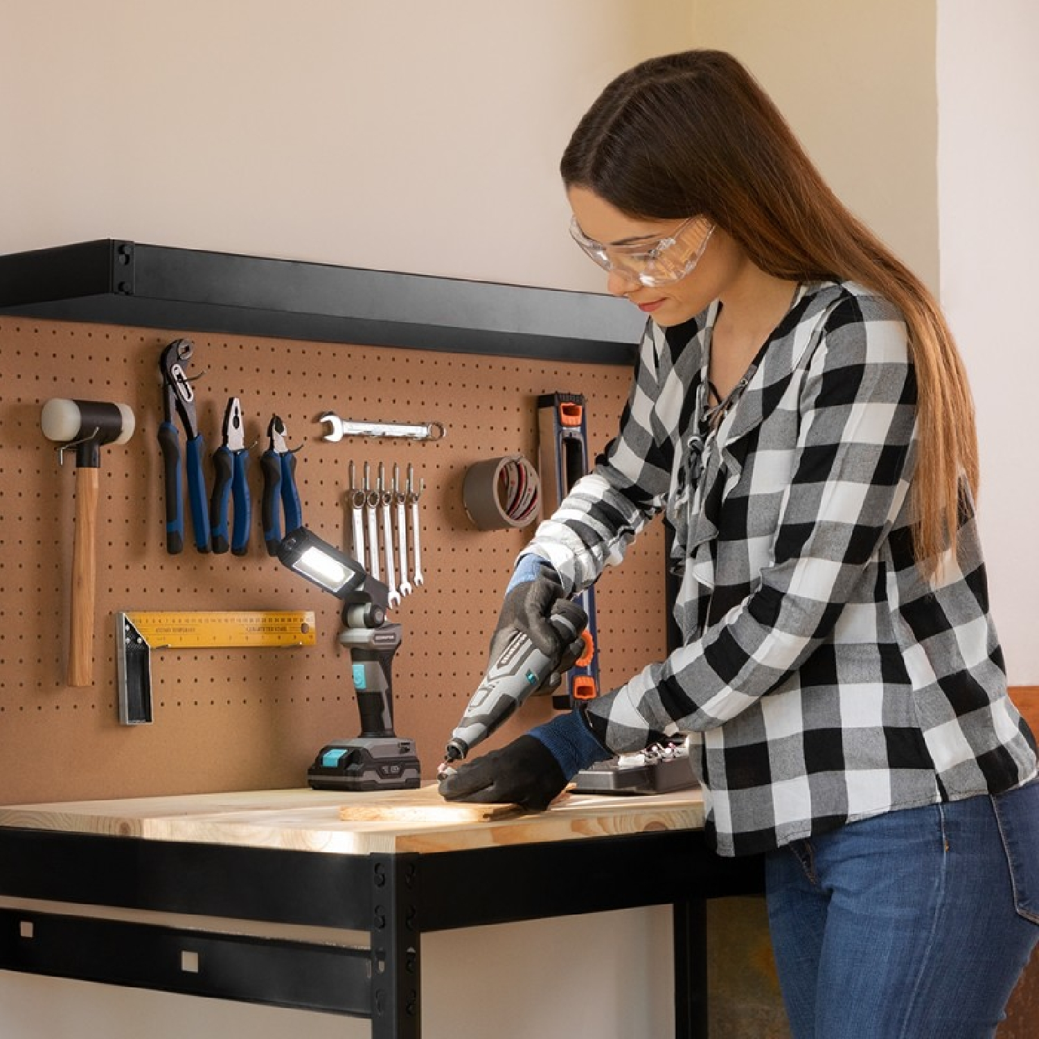 Kobieta używa narzędzia obrotowego przy stole warsztatowym. Nosi okulary ochronne i rękawice, pracuje z drewnem.