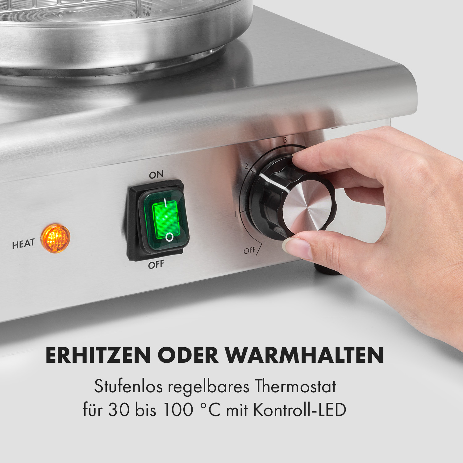 Eine Hand dreht einen schwarzen Knopf an einem silbernen Gerät, mit einem grünen Ein/Aus-Schalter und orangefarbenem Heizlicht.