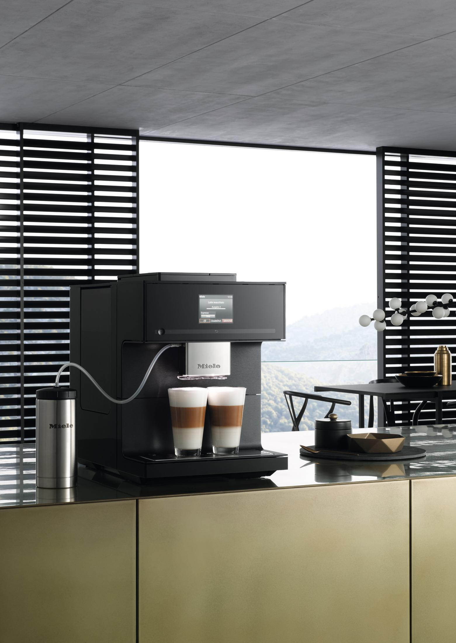 Schwarze Kaffeemaschine auf goldener Theke, zwei Latte Macchiato, Bergpanorama im Hintergrund.