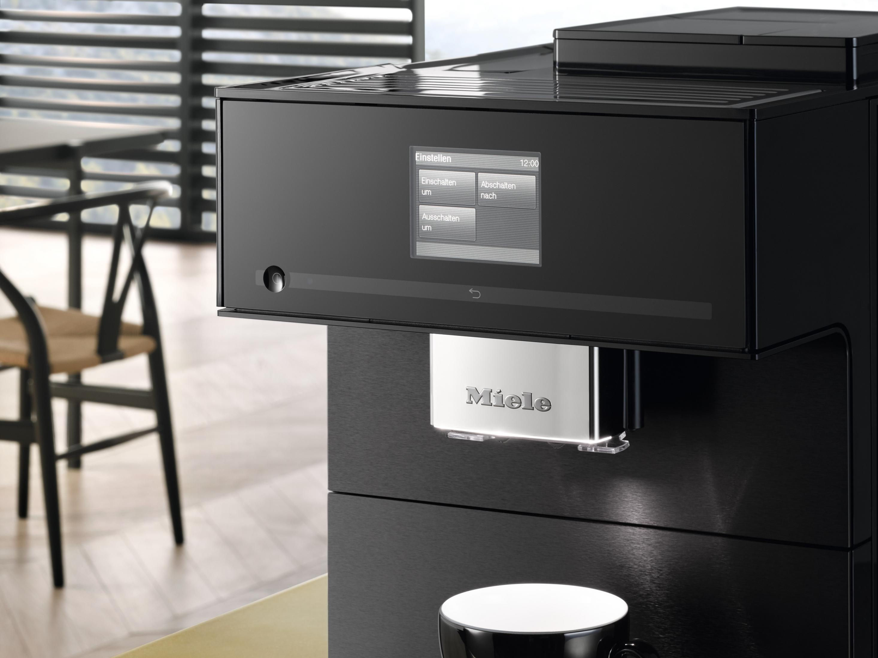 Schwarze Kaffeemaschine mit Bildschirm, der Einstellungen zeigt. Unter dem Auslauf steht eine Tasse, ein Stuhl im Hintergrund.