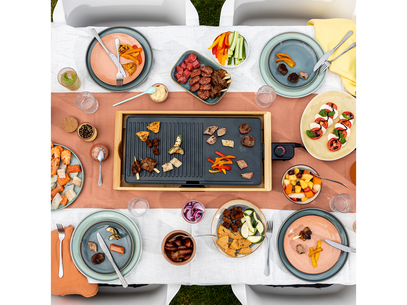 Tisch mit Grill, Essen, Tellern und Getränken. Sitzgelegenheit im Freien mit weißen Stühlen.