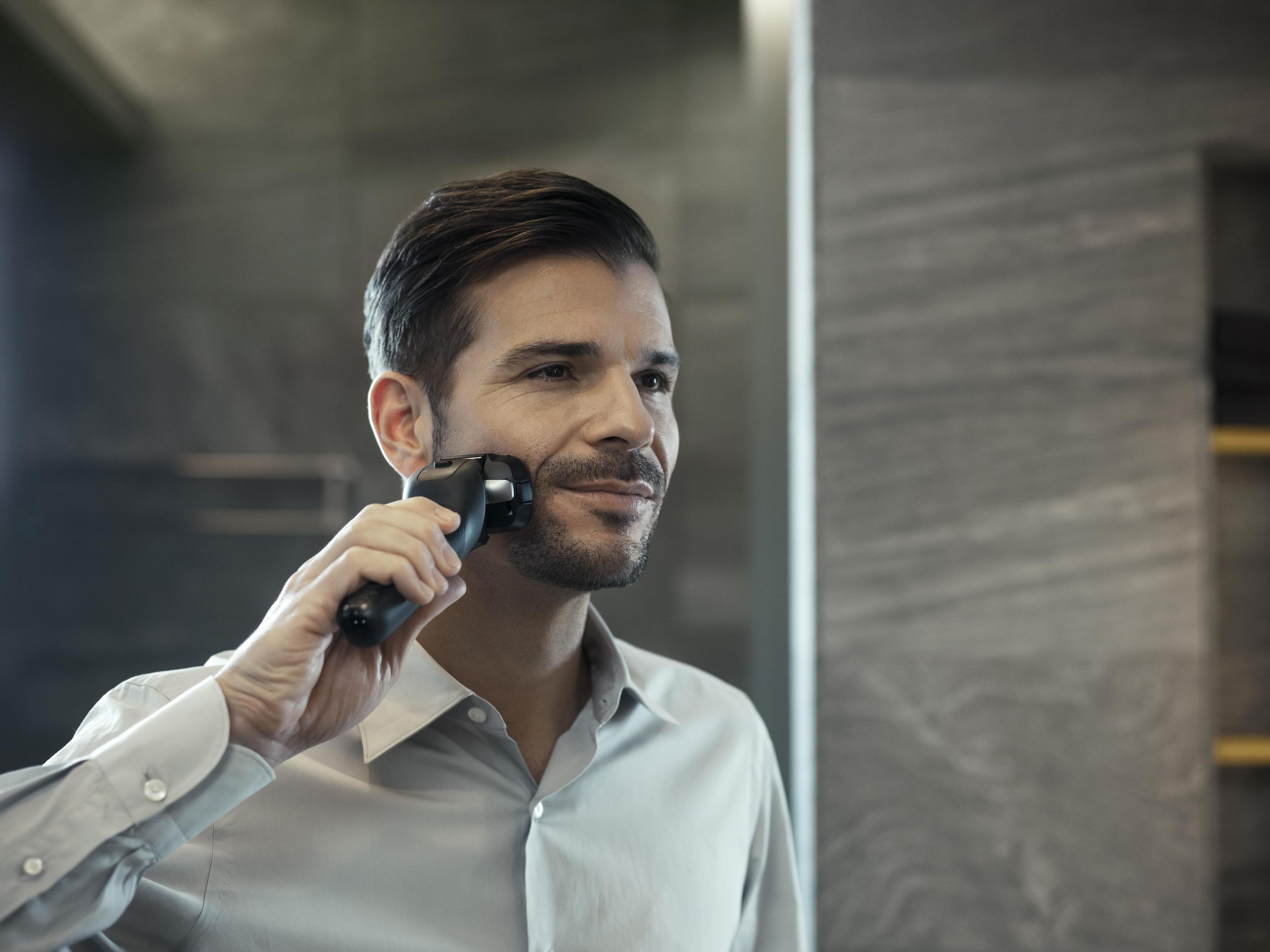 Mężczyzna goli twarz golarką elektryczną, patrząc w lustro.