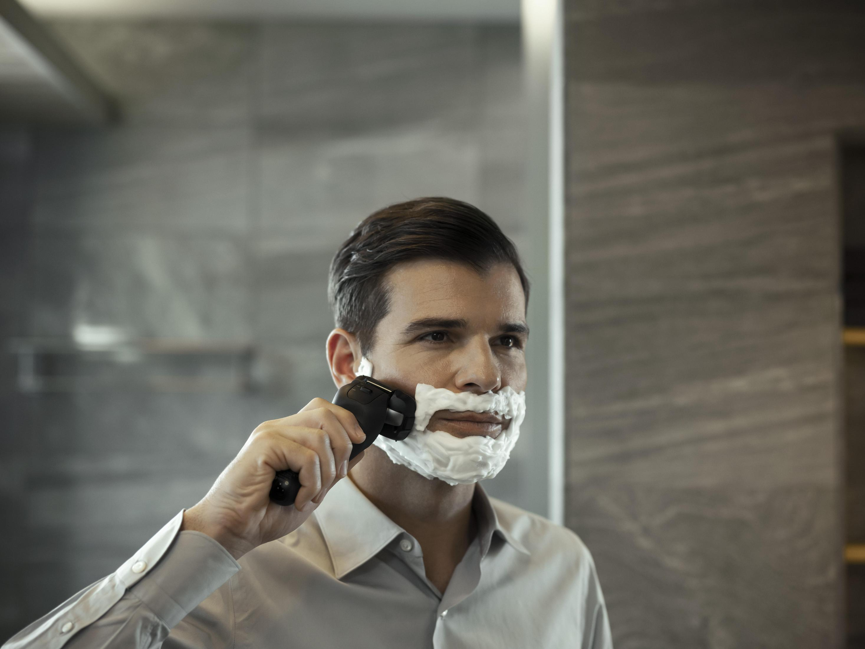 Mężczyzna goli się golarką elektryczną. Piana jest nałożona na jego twarz. Lustro jest za nim.