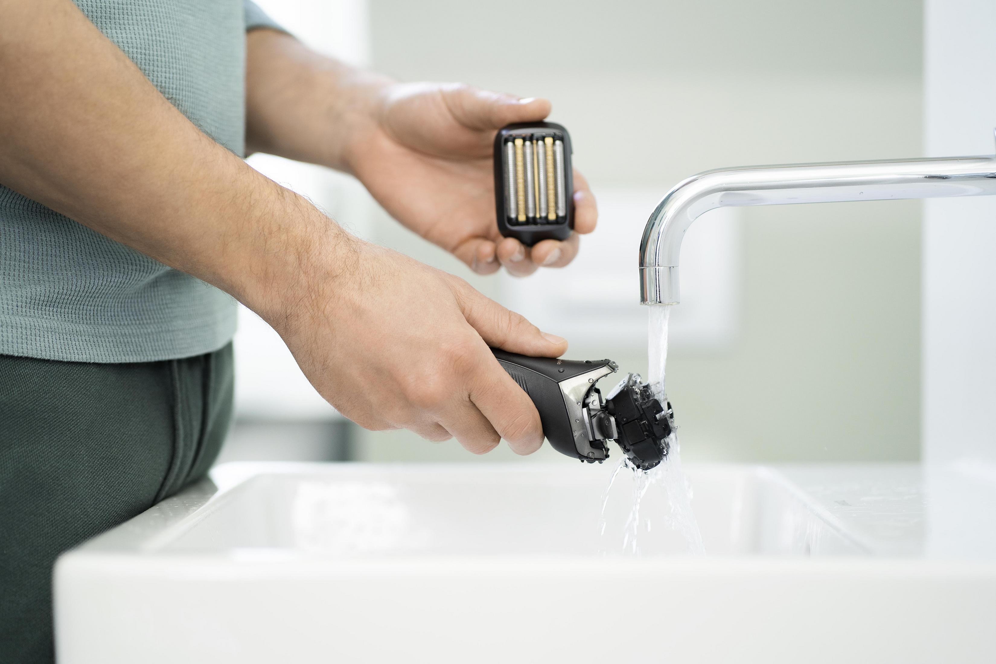 Mężczyzna myjący głowicę golarki pod kranem. Trzyma golarkę i głowicę zamienną.
