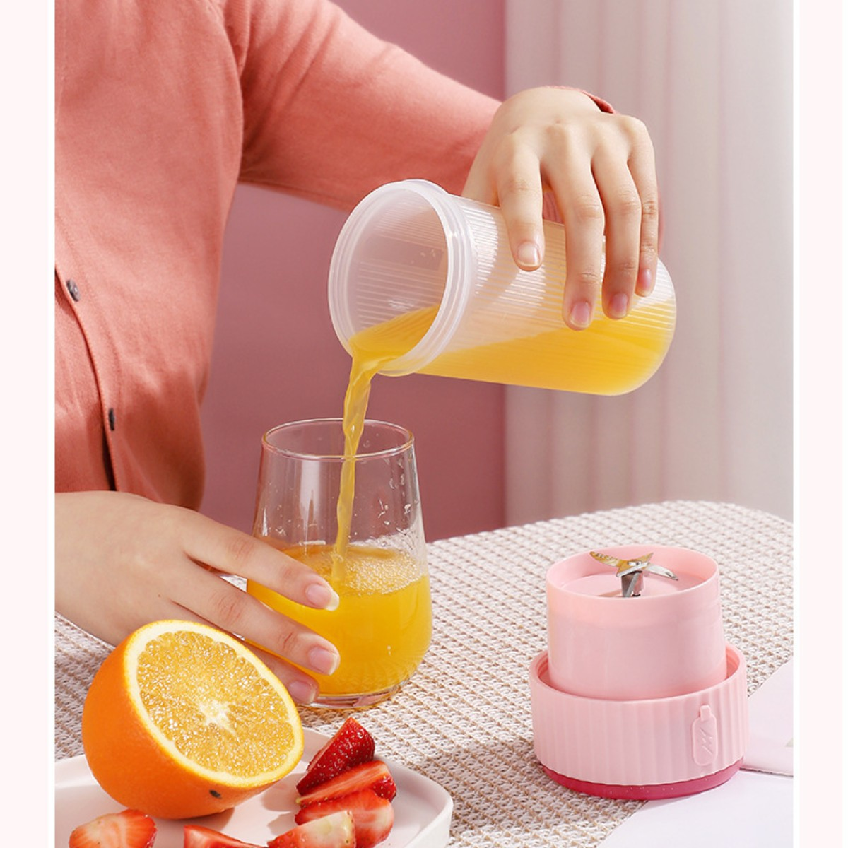 Eine Person gießt Orangensaft aus einem Behälter in ein Glas. Es gibt einen Mixer, eine Orange und Erdbeeren.
