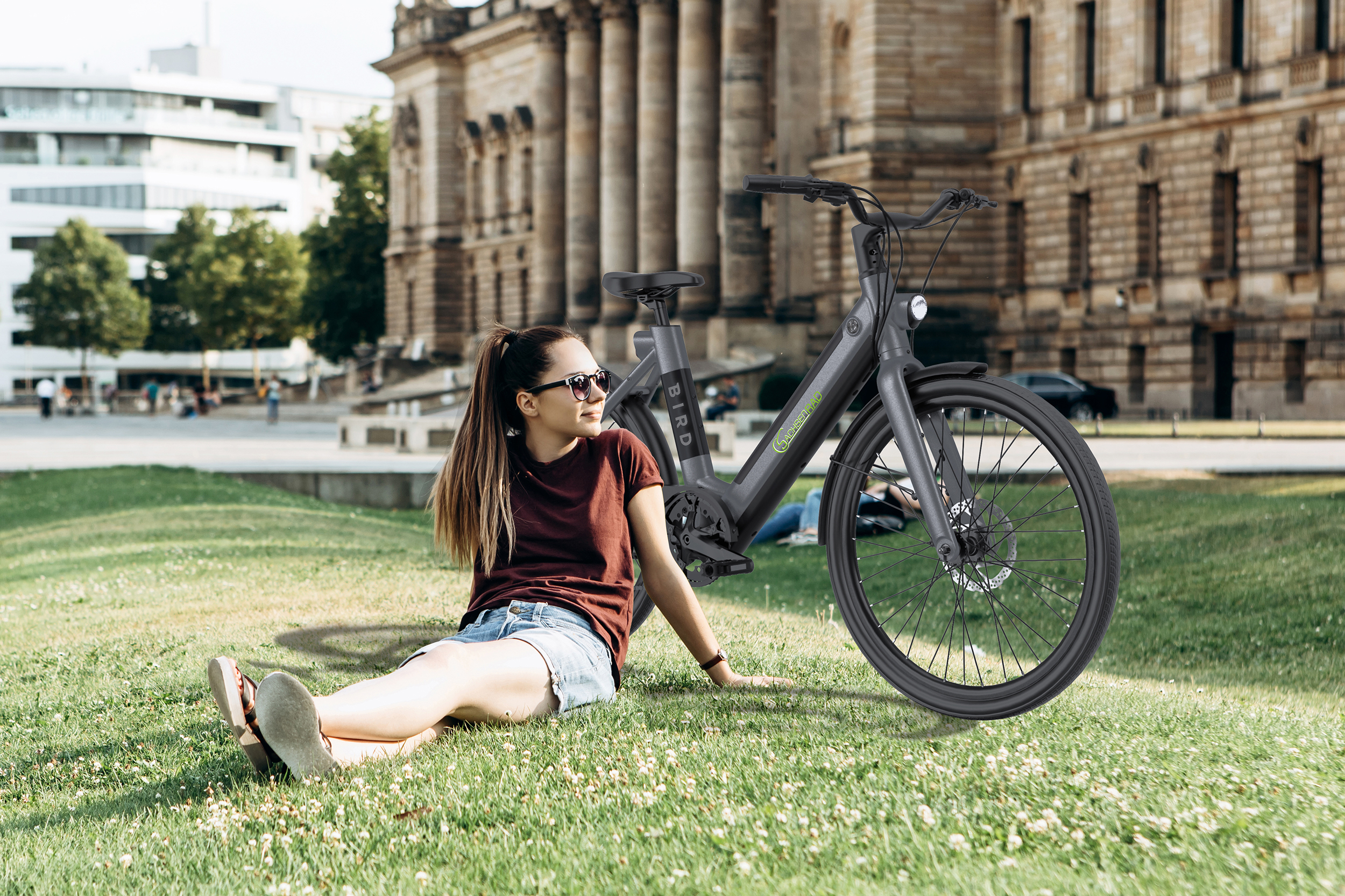 Frau auf Gras mit Fahrrad. Das Fahrrad ist grau. Im Hintergrund ein Gebäude und Bäume.