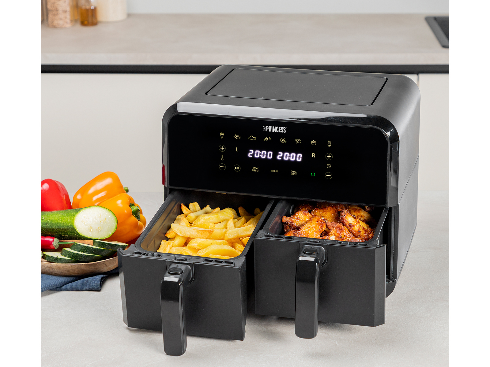 Czarna frytkownica powietrzna z dwoma przegrodami, jedna z frytkami, druga ze skrzydełkami kurczaka, warzywa w tle.