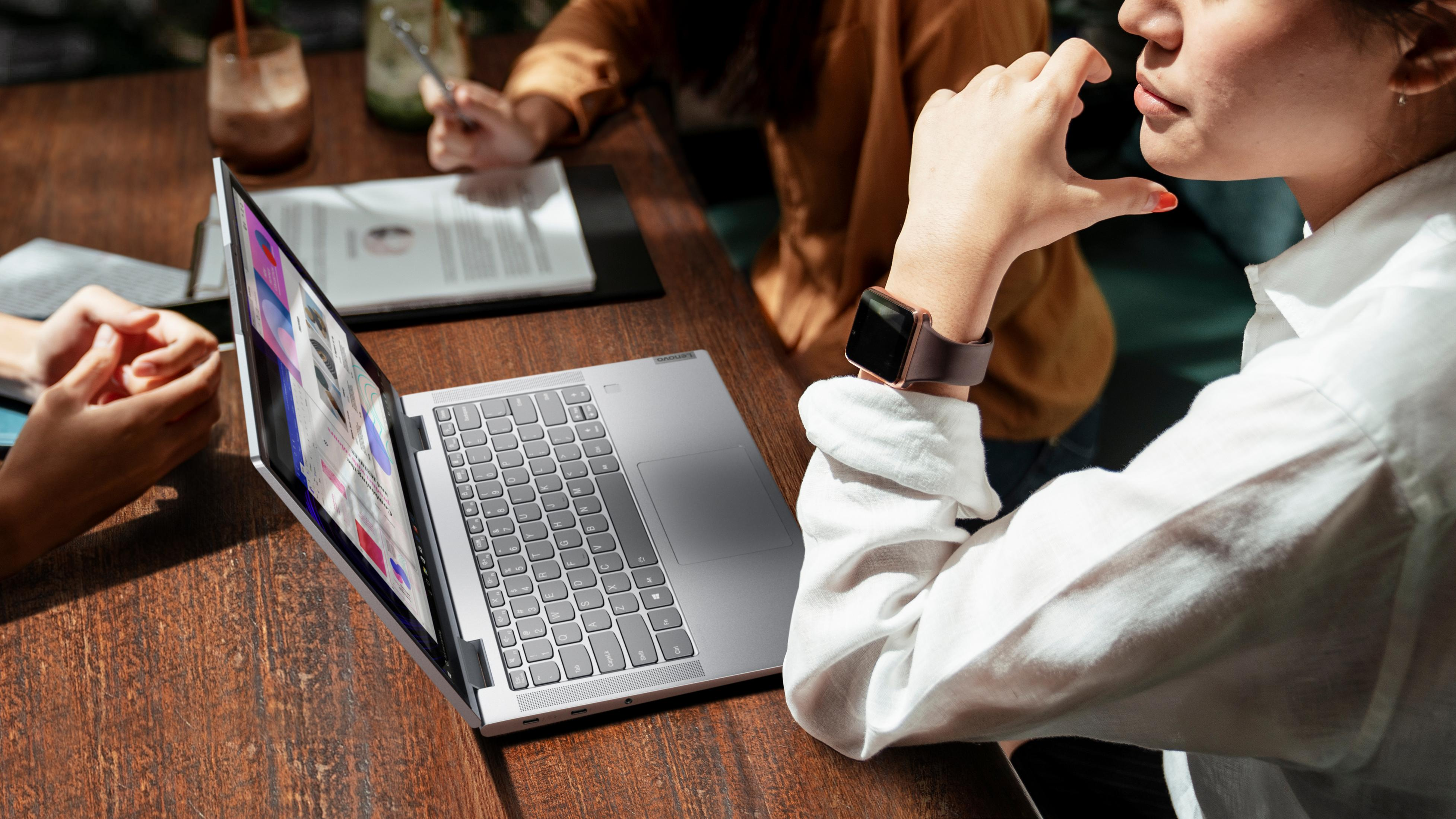 Eine Frau mit Smartwatch benutzt einen Laptop. Zwei weitere Personen im Hintergrund am Tisch.